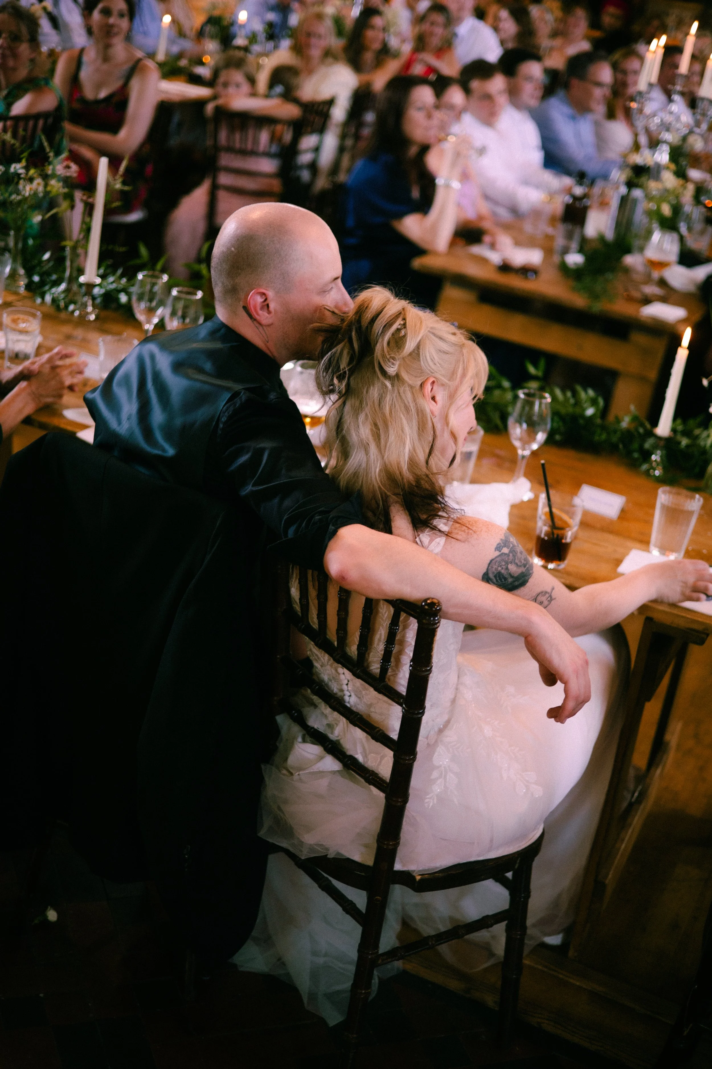 A wedding reception with a newlywed couple sitting at a table, the groom with a shaved head and the bride with blonde hair and tattoos, smiling and enjoying the celebration, surrounded by friends and family in a warmly lit room with candles and flora