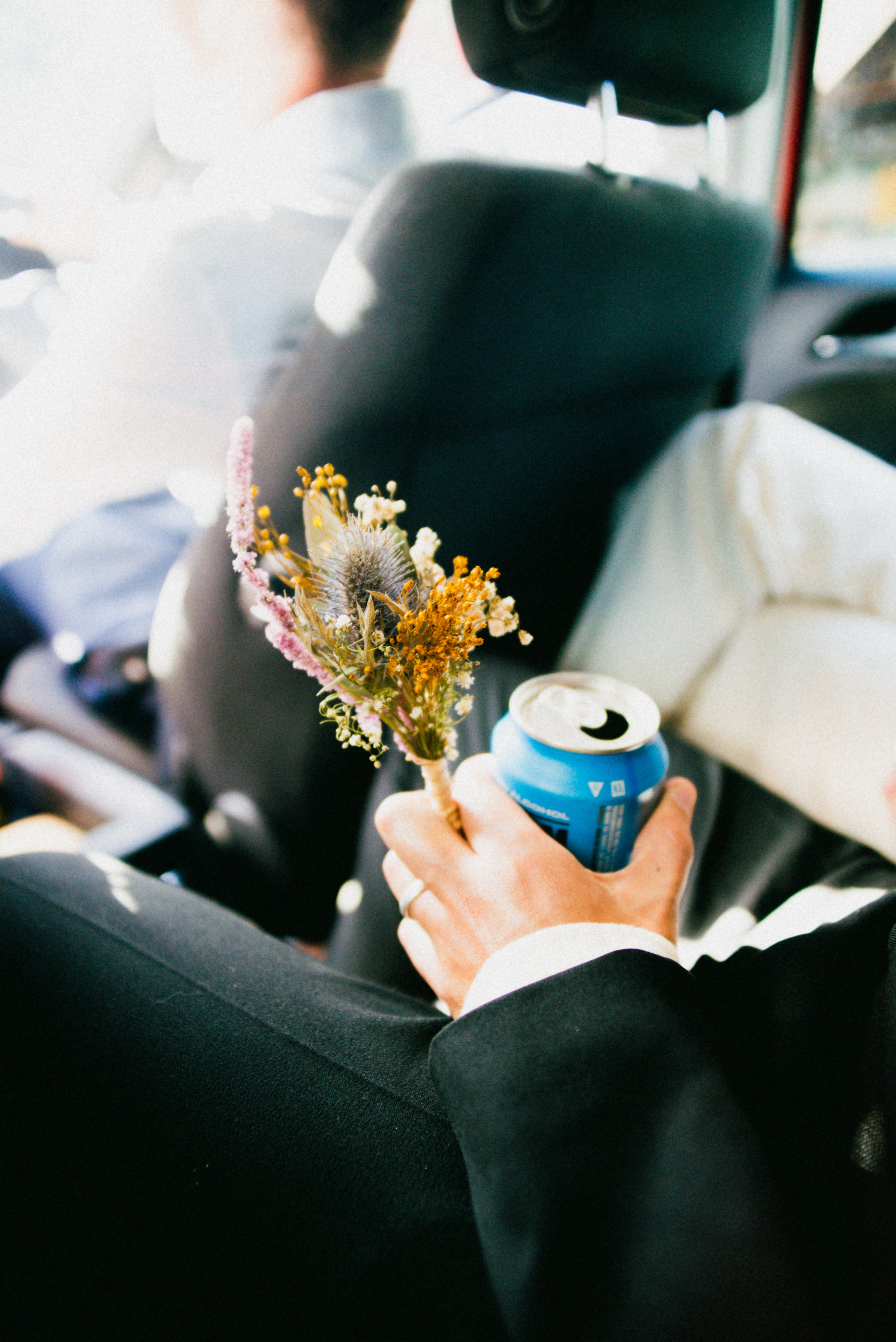 Person sitting in a car holding a small bouquet of dried wildflowers and an open can of Bud Light beer.