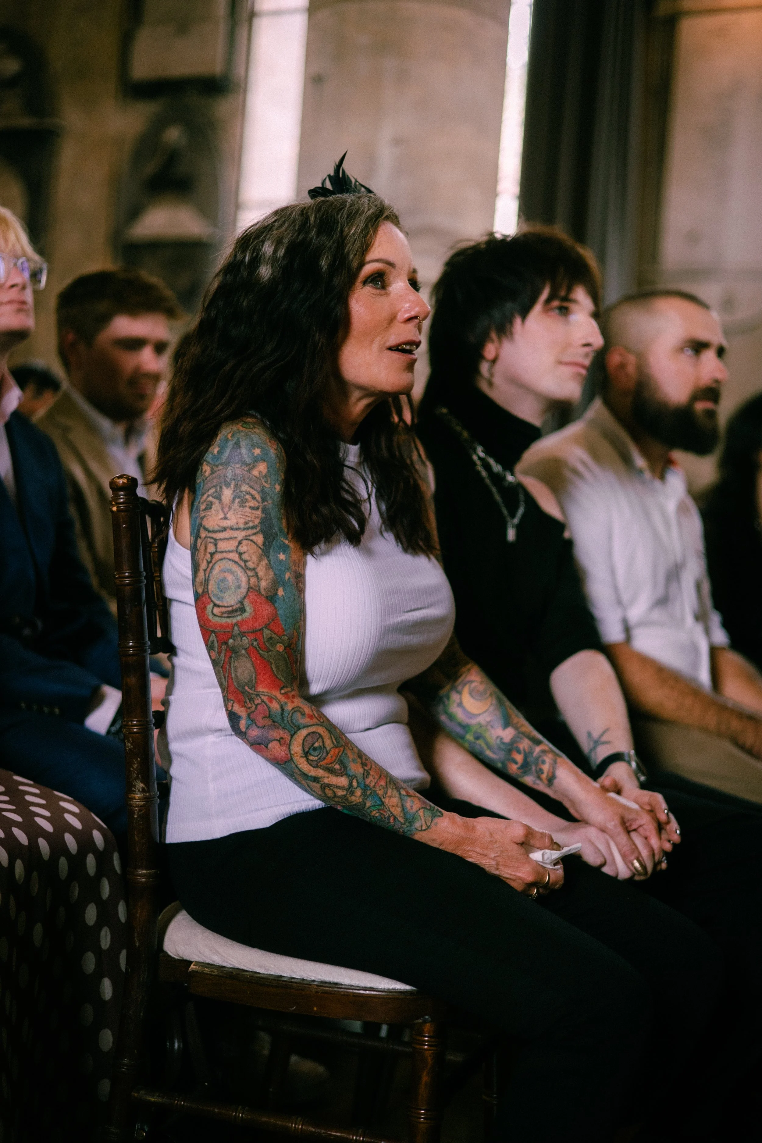 A group of five people sitting in a row at an indoor event. The woman in the foreground has long dark wavy hair, multiple colorful tattoos on her arms, and is wearing a white sleeveless top. She appears to be speaking or reacting to something. The ot