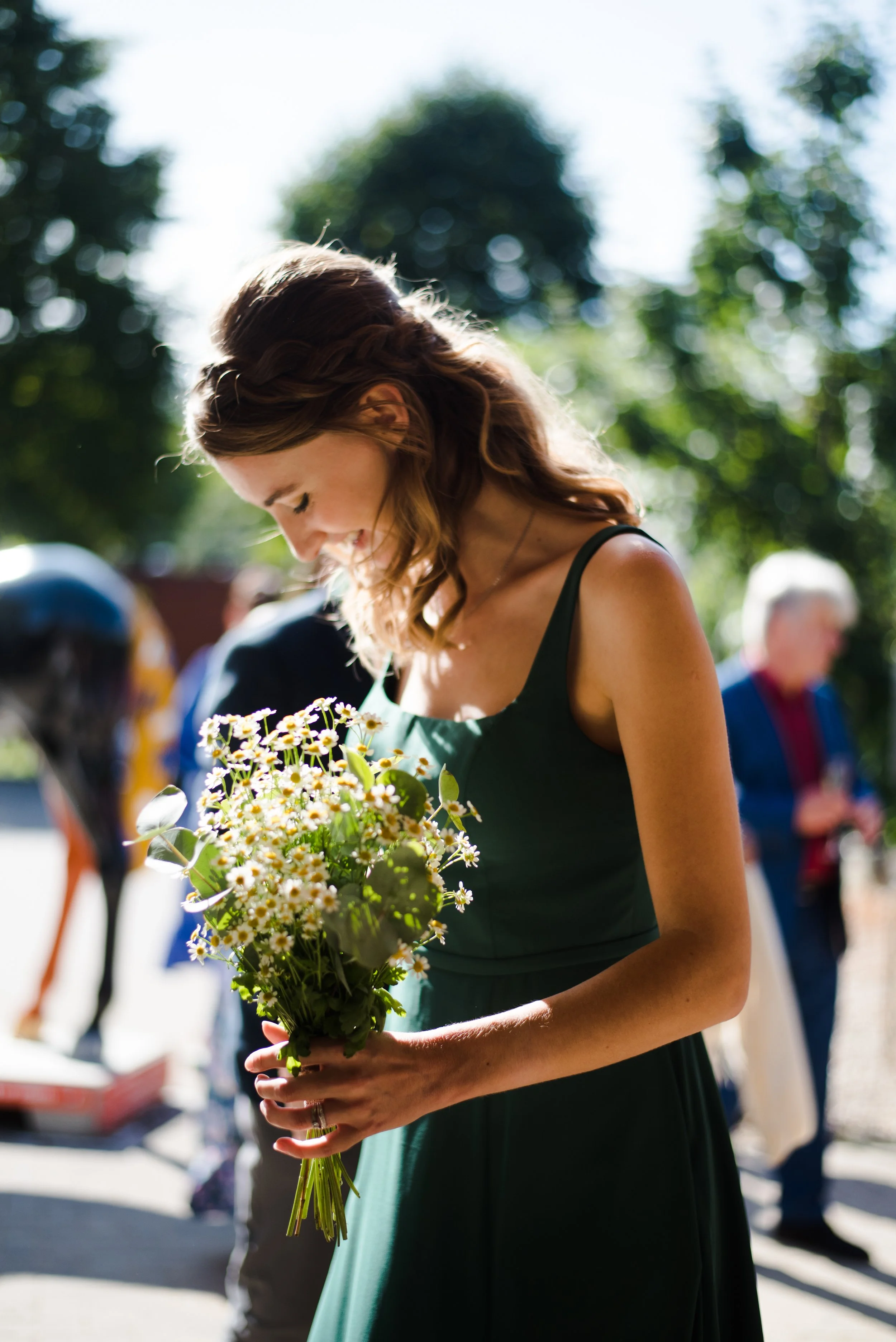 A young woman in a green dress holding a bouquet of daisies, smiling with her head bowed, outdoors in sunlight.