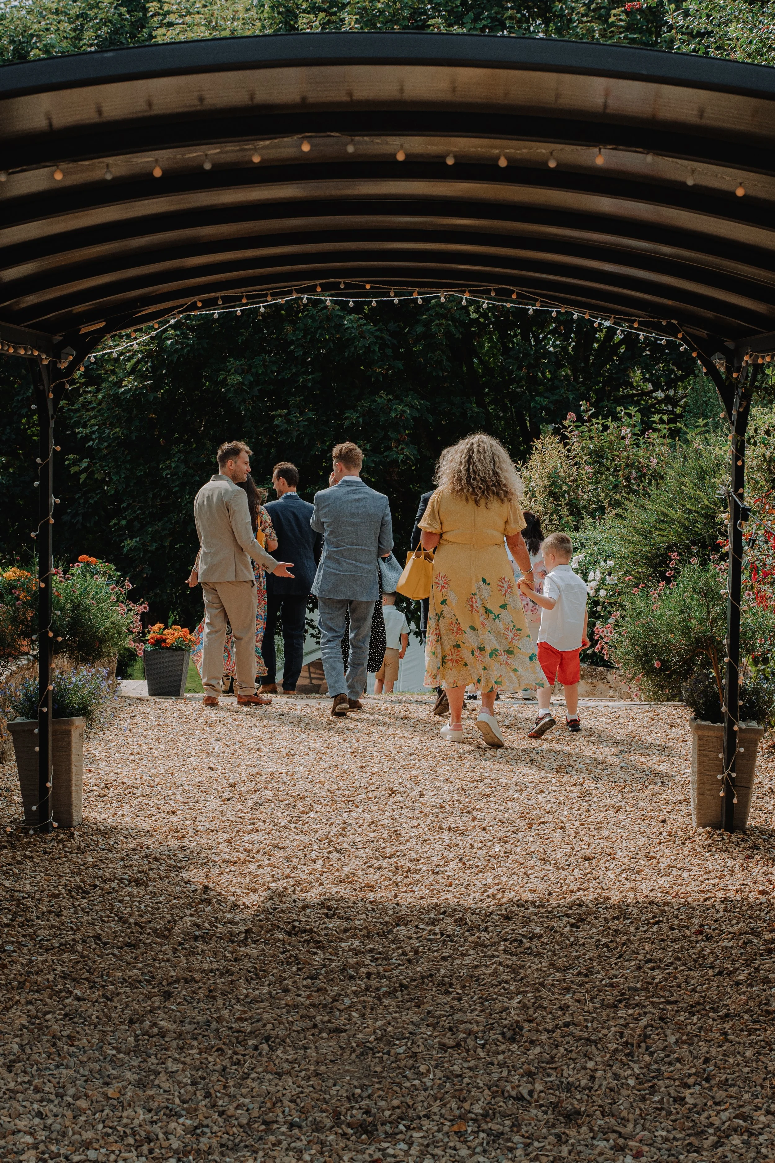 Group of people walking under a garden arbor decorated with string lights, surrounded by potted flowers.