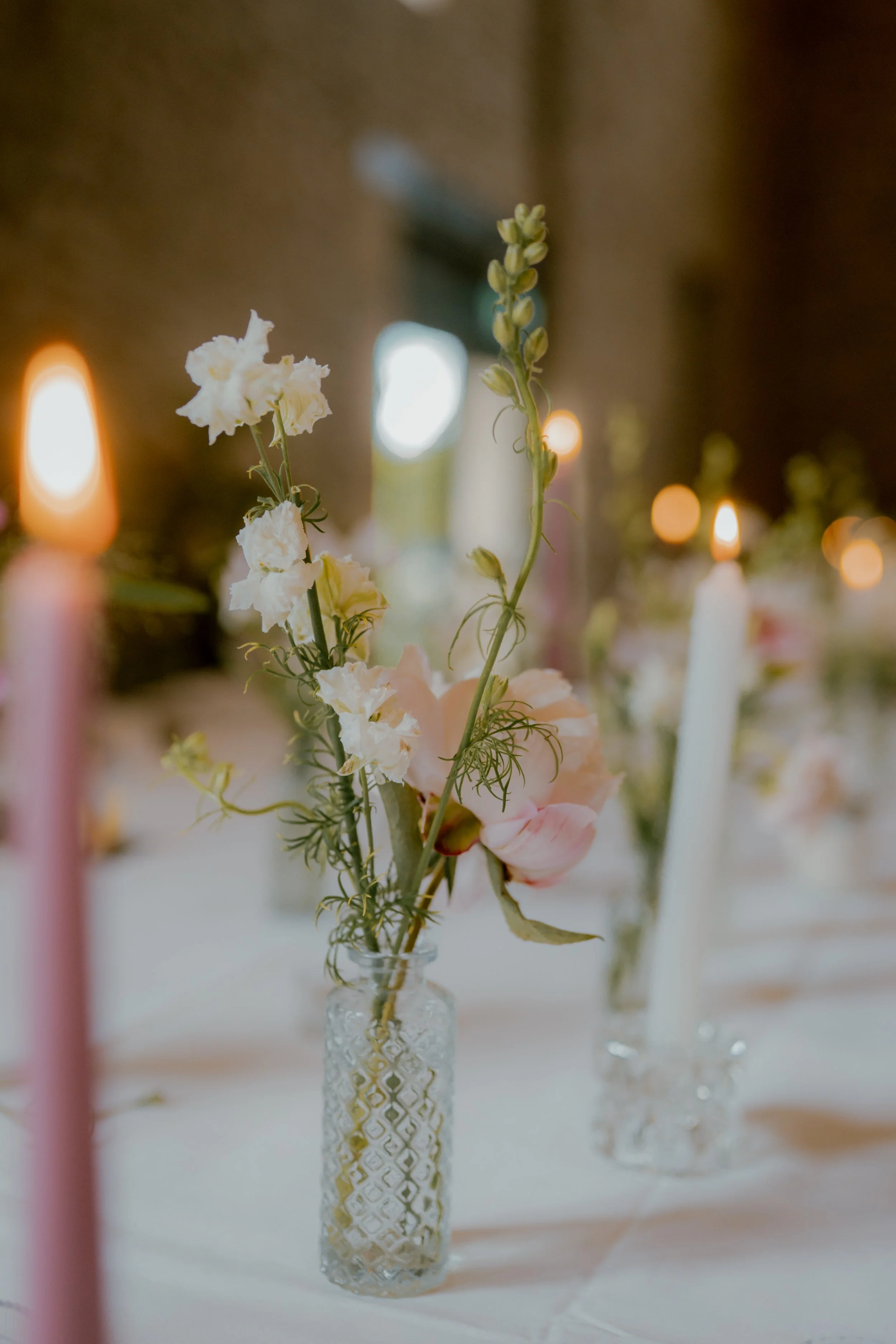 A floral arrangement in a textured glass vase on a table with lit candles and blurred background.