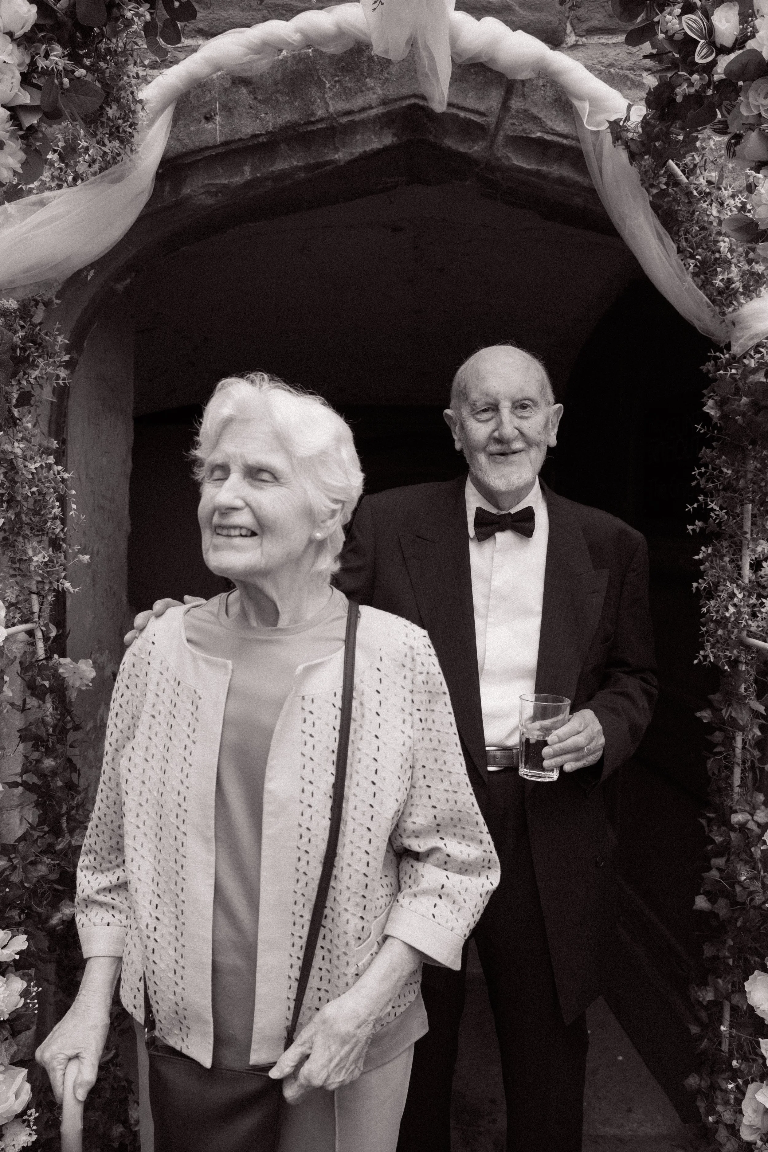 An elderly couple smiling and standing together at a celebration, framed by an arch decorated with flowers and draped fabric.