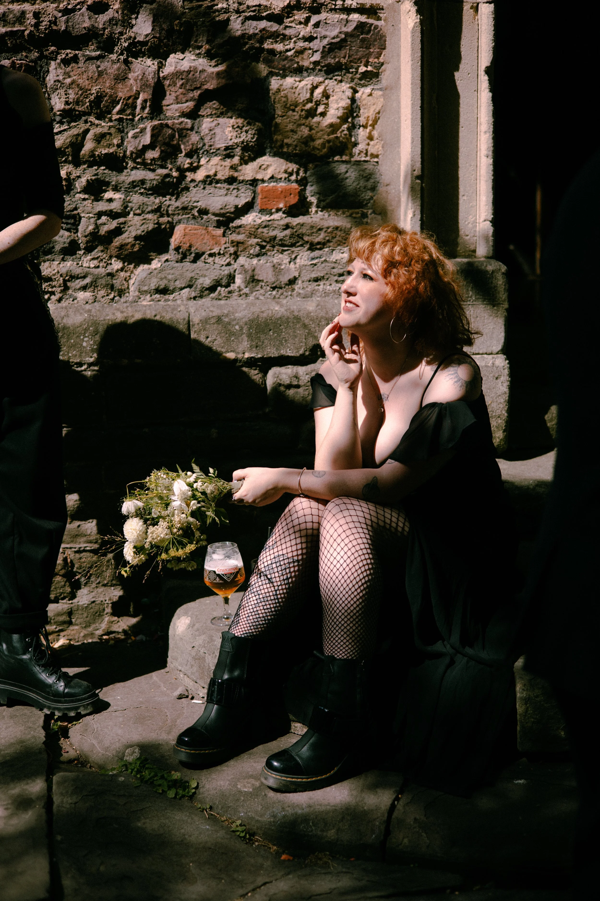 A woman with curly red hair sitting by a stone wall, wearing a black dress, fishnet stockings, and black boots. She rests her chin on her hand and looks thoughtfully into the distance. A bouquet of white flowers and a glass of beer are placed on the 