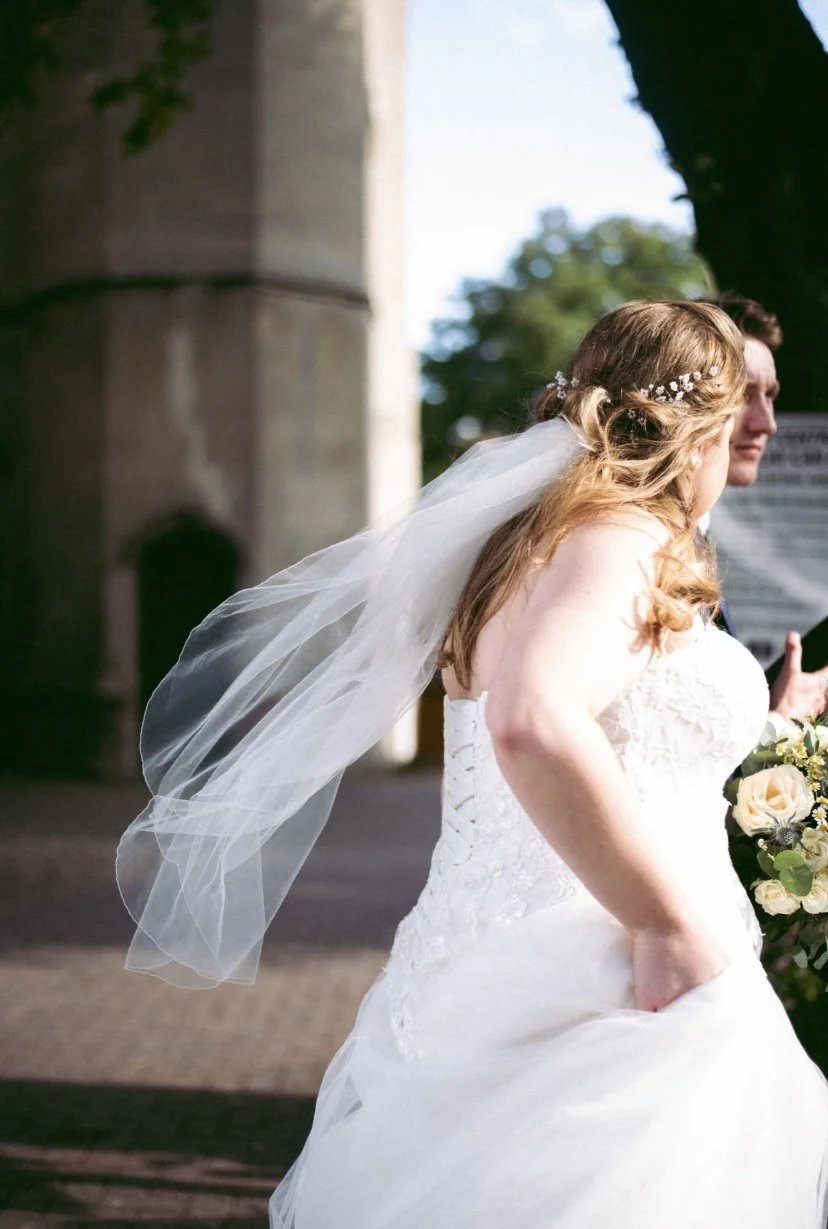 A bride with long, wavy, light brown hair wearing a white strapless wedding gown, a veil, and a floral headband, holding a bouquet of white and yellow roses, standing outdoors.
