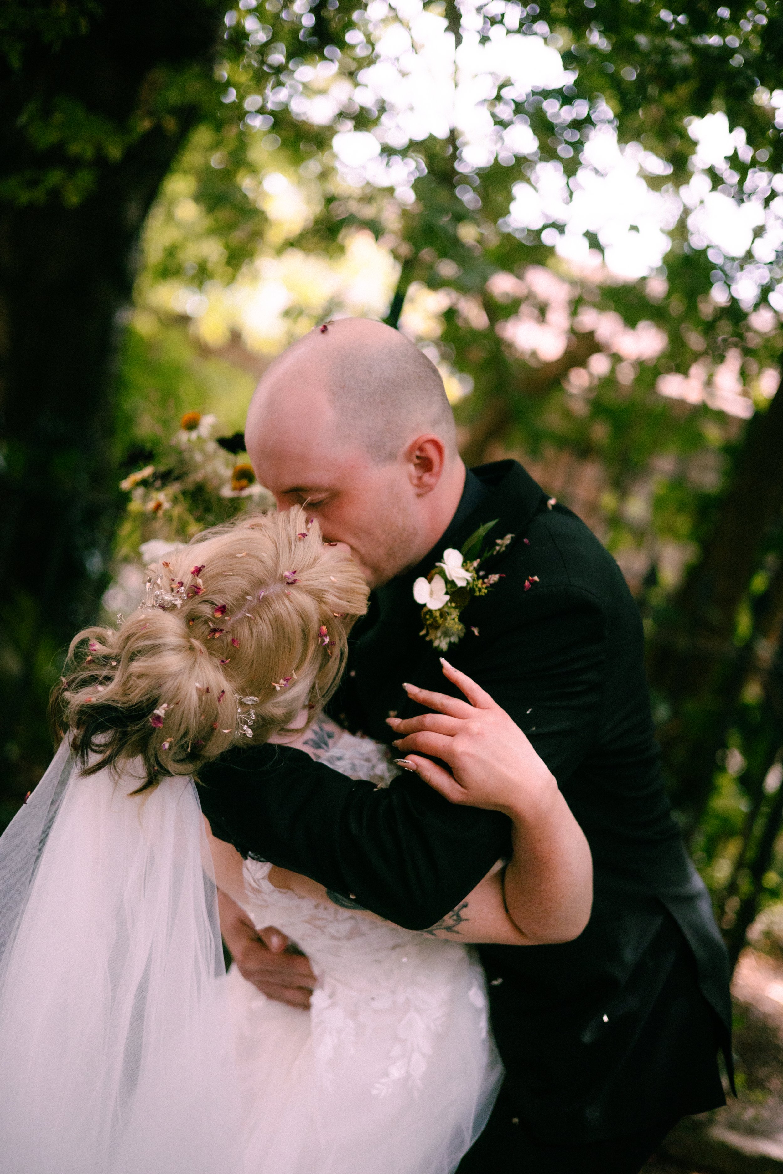 A couple sharing a kiss during a wedding ceremony outdoors with green trees and sunlight in the background. The bride has curly blonde hair with flower decorations and is wearing a white wedding gown. The groom has a shaved head, black suit, and a bo