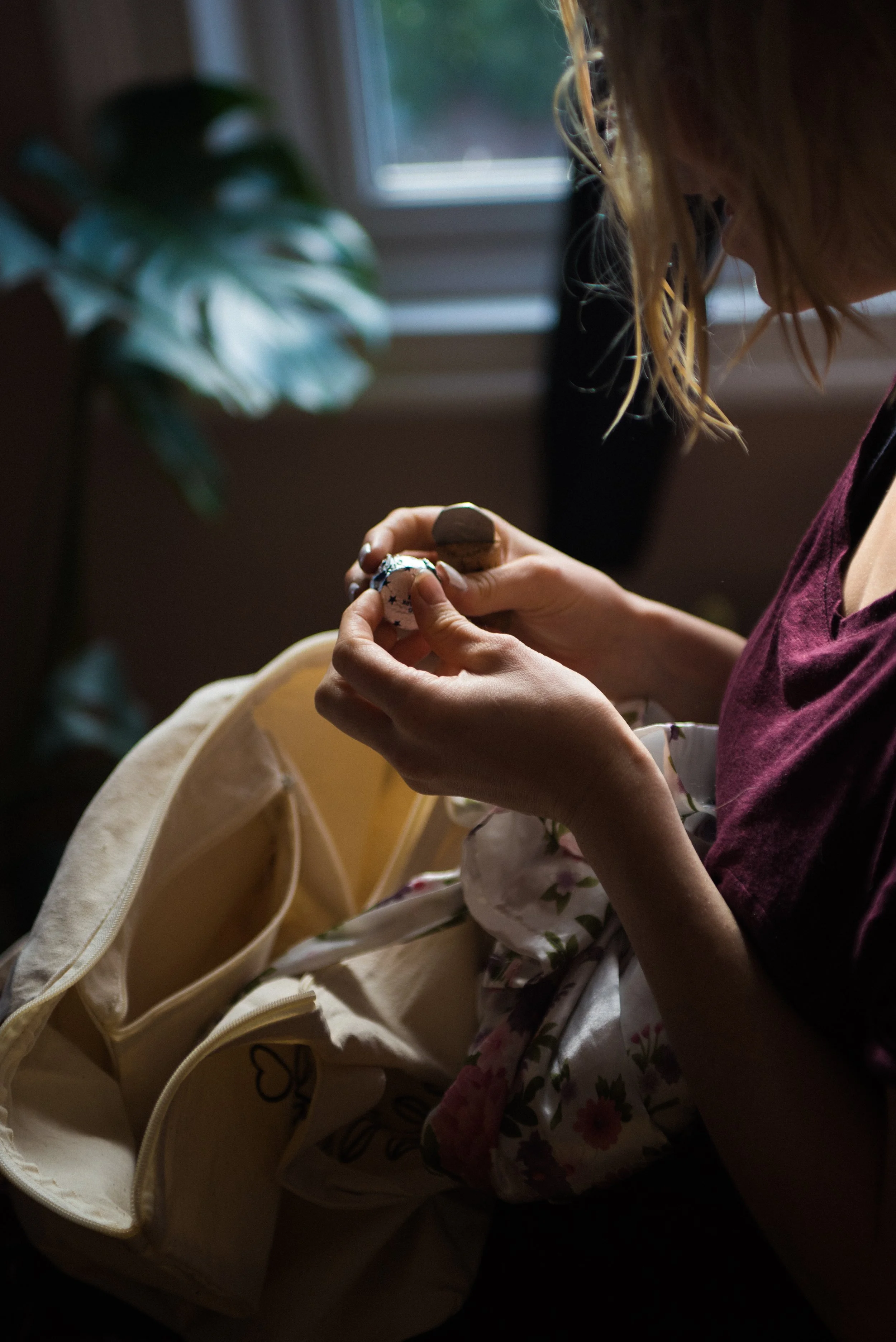 A woman with blonde hair, wearing a maroon top, is sitting in a room near a window, holding a small object and a cork, with an open cream-colored bag on her lap.