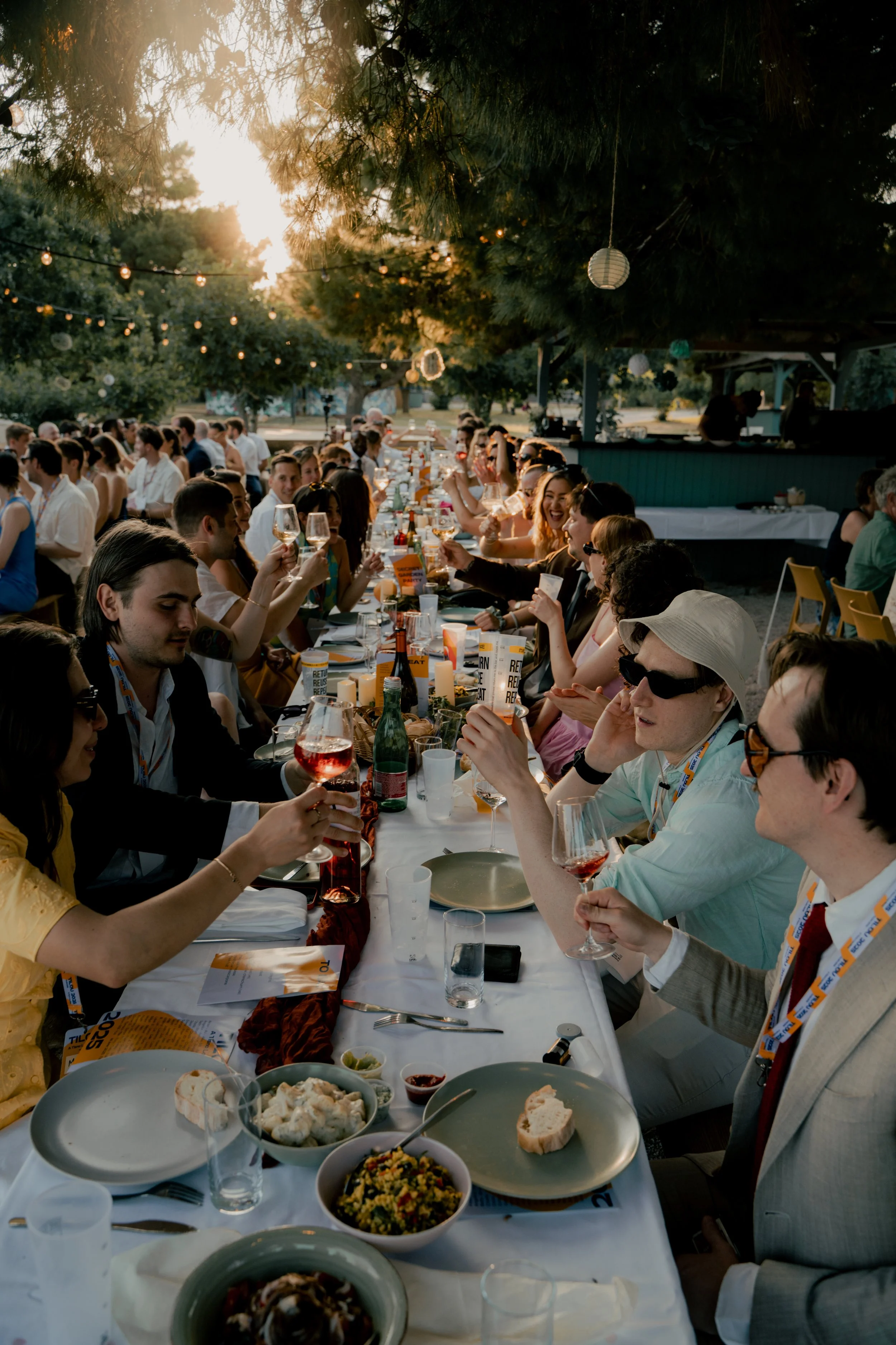 People dining at a long outdoor table with drinks and food during sunset, surrounded by string lights and trees.