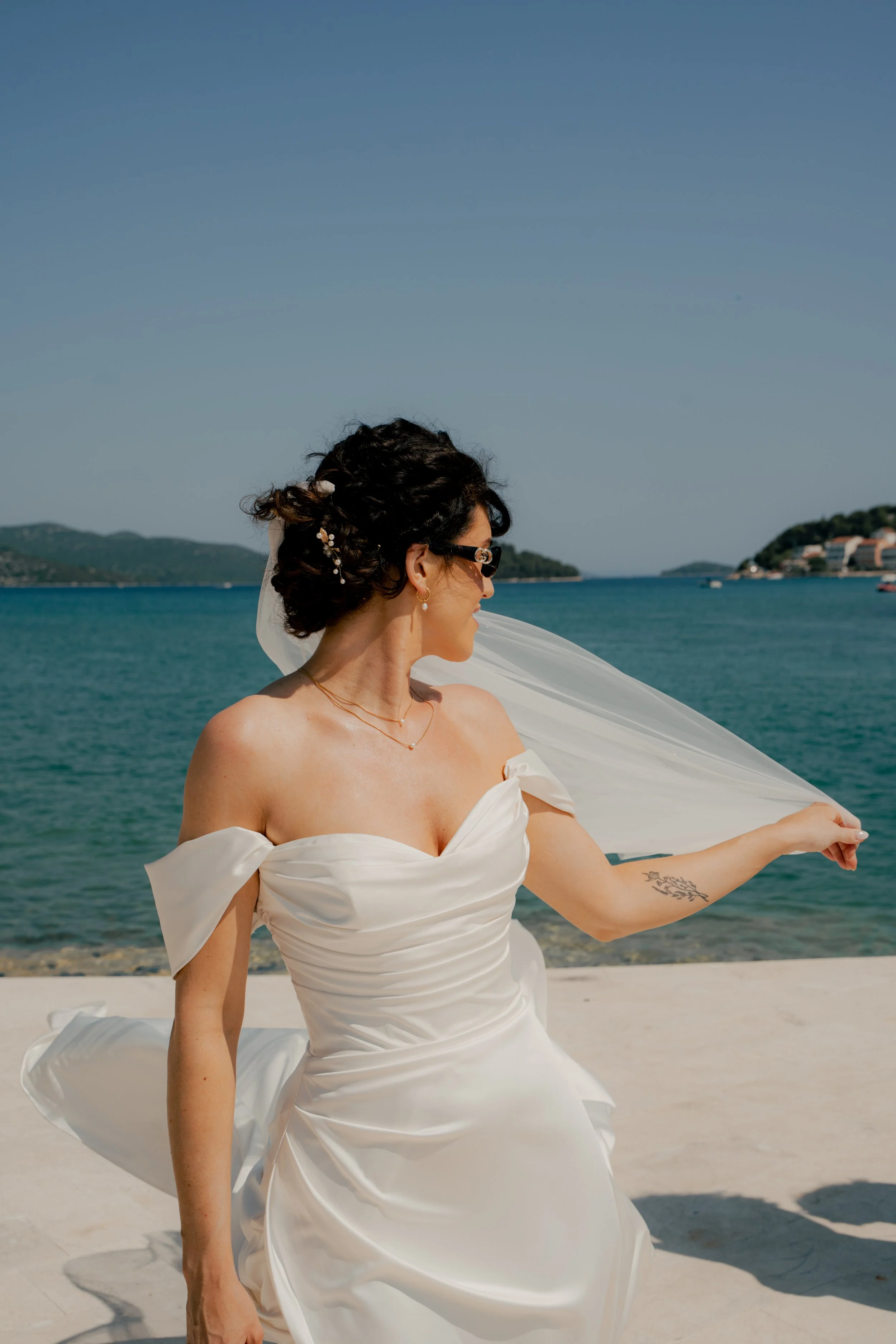 Bride in a white wedding gown holding a veil near her face by the water with boats and hills in the background.
