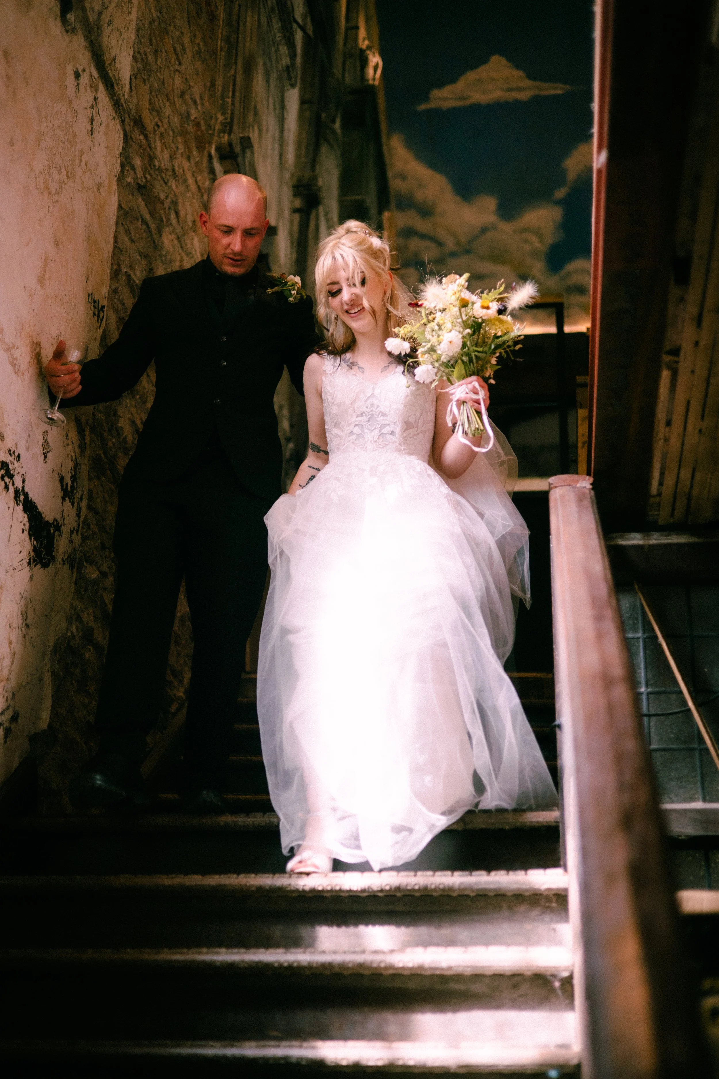 A bride in a white wedding gown holding a bouquet of flowers and smiling, with a groom in a black suit and holding a glass, descending a rustic staircase with a mural of clouds on the wall behind them.