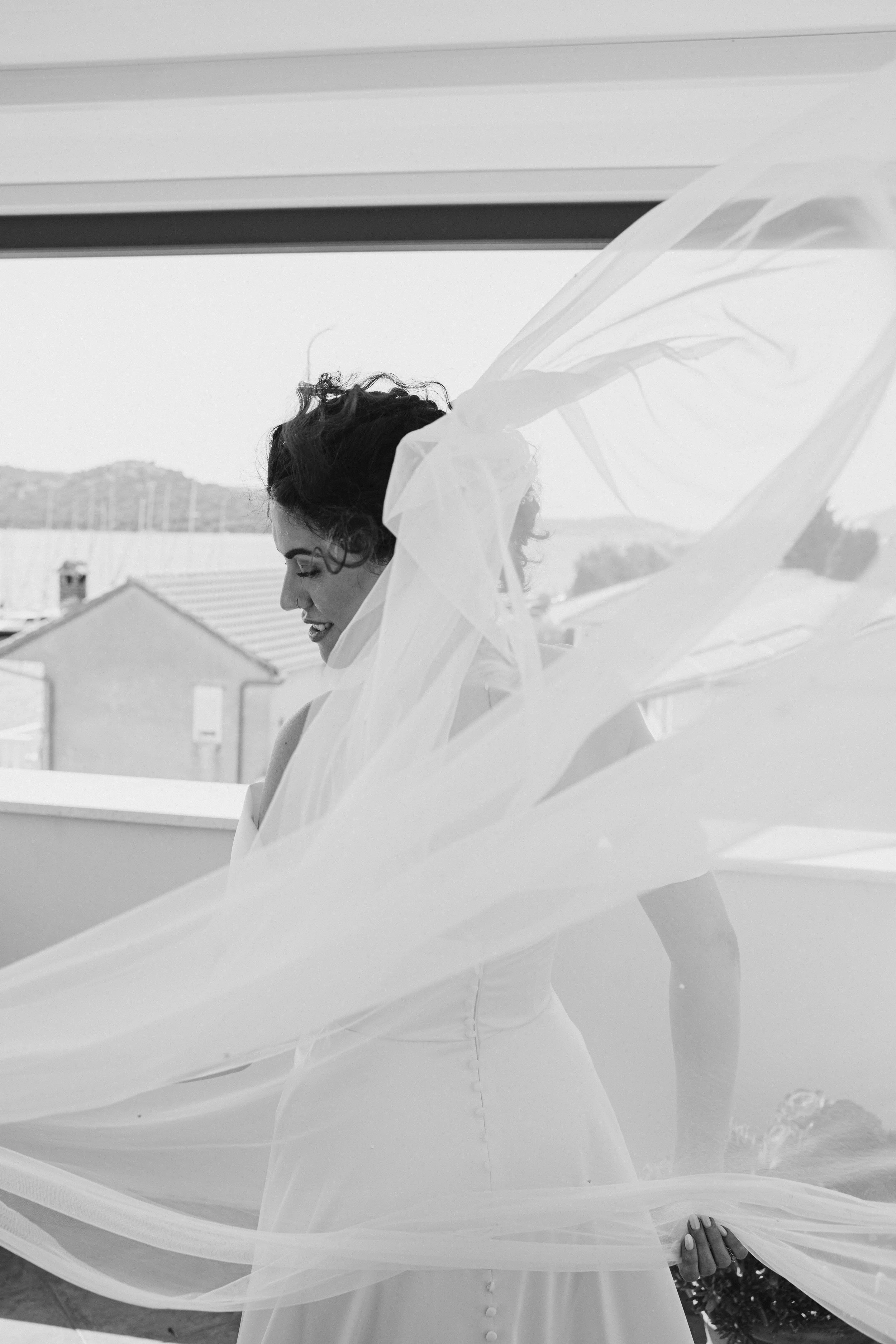 A woman in a wedding dress smiling with a flowing veil, standing near a window with a view of houses and hills in the background.