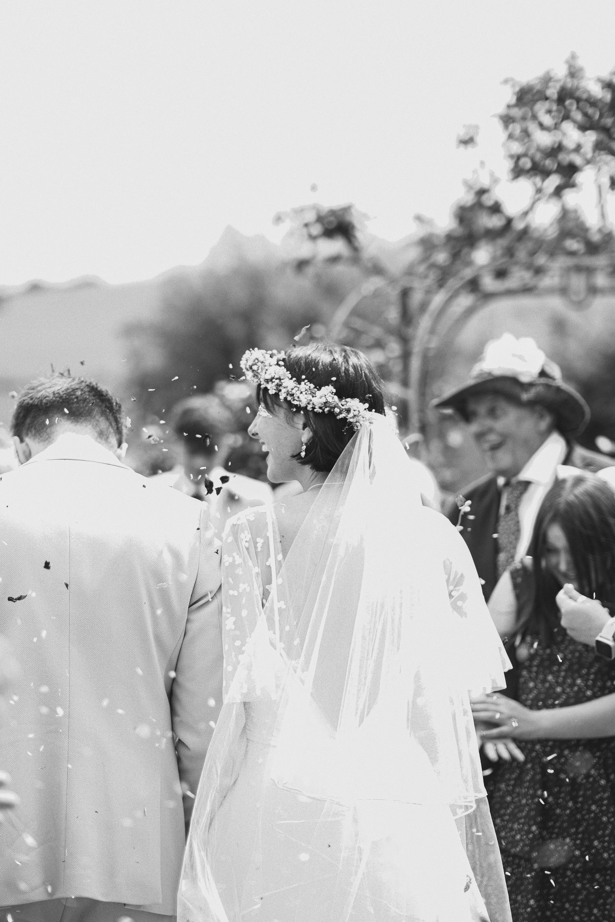 Black and white photo of a bride wearing a floral crown and veil, smiling during a wedding celebration outdoors, surrounded by guests.