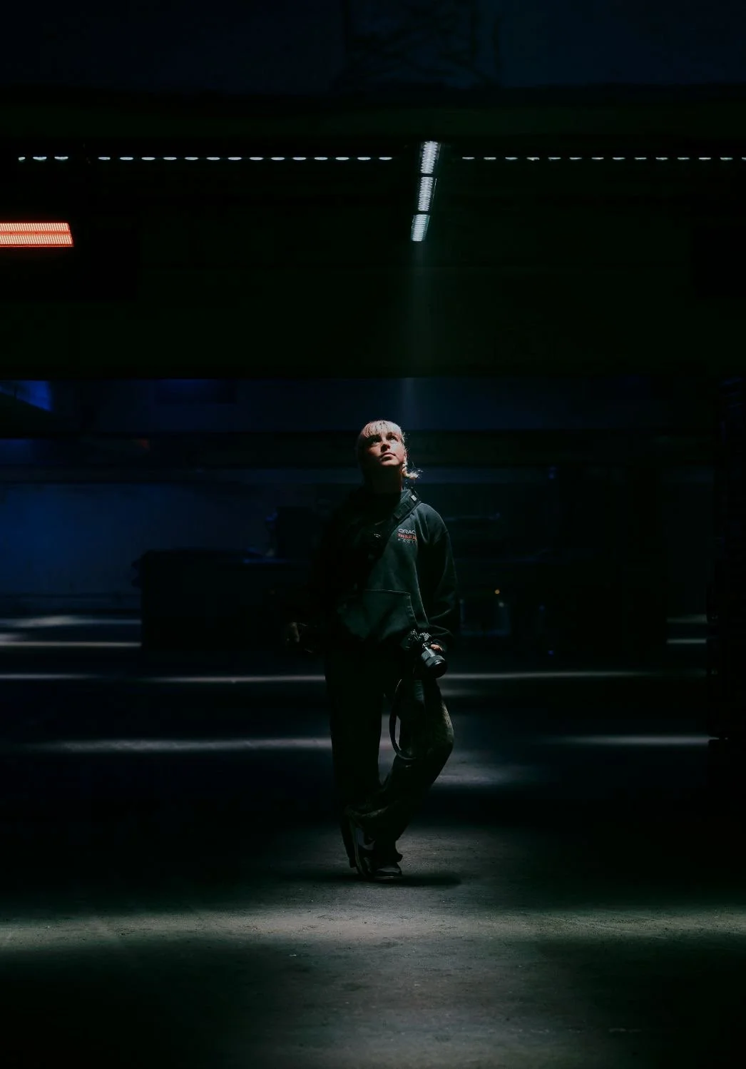 Person standing in a dimly lit indoor parking garage, holding a camera, looking up toward light coming from above.