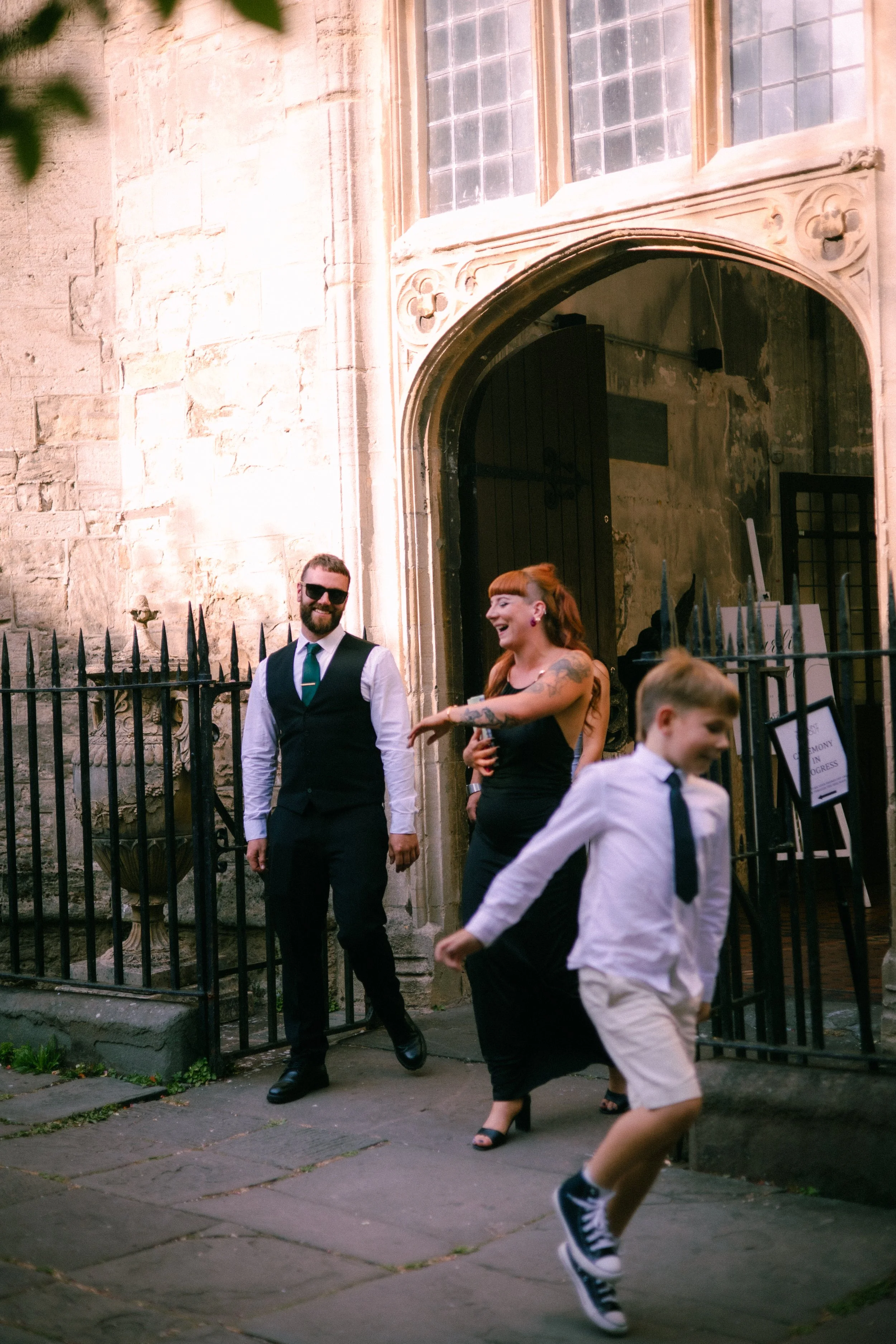 A man in a black vest, white shirt, blue tie, and black sunglasses stands smiling near a black wrought-iron fence outside a stone building with a large arched doorway. A woman with red hair, black dress, and high heels is laughing and reaching out. A