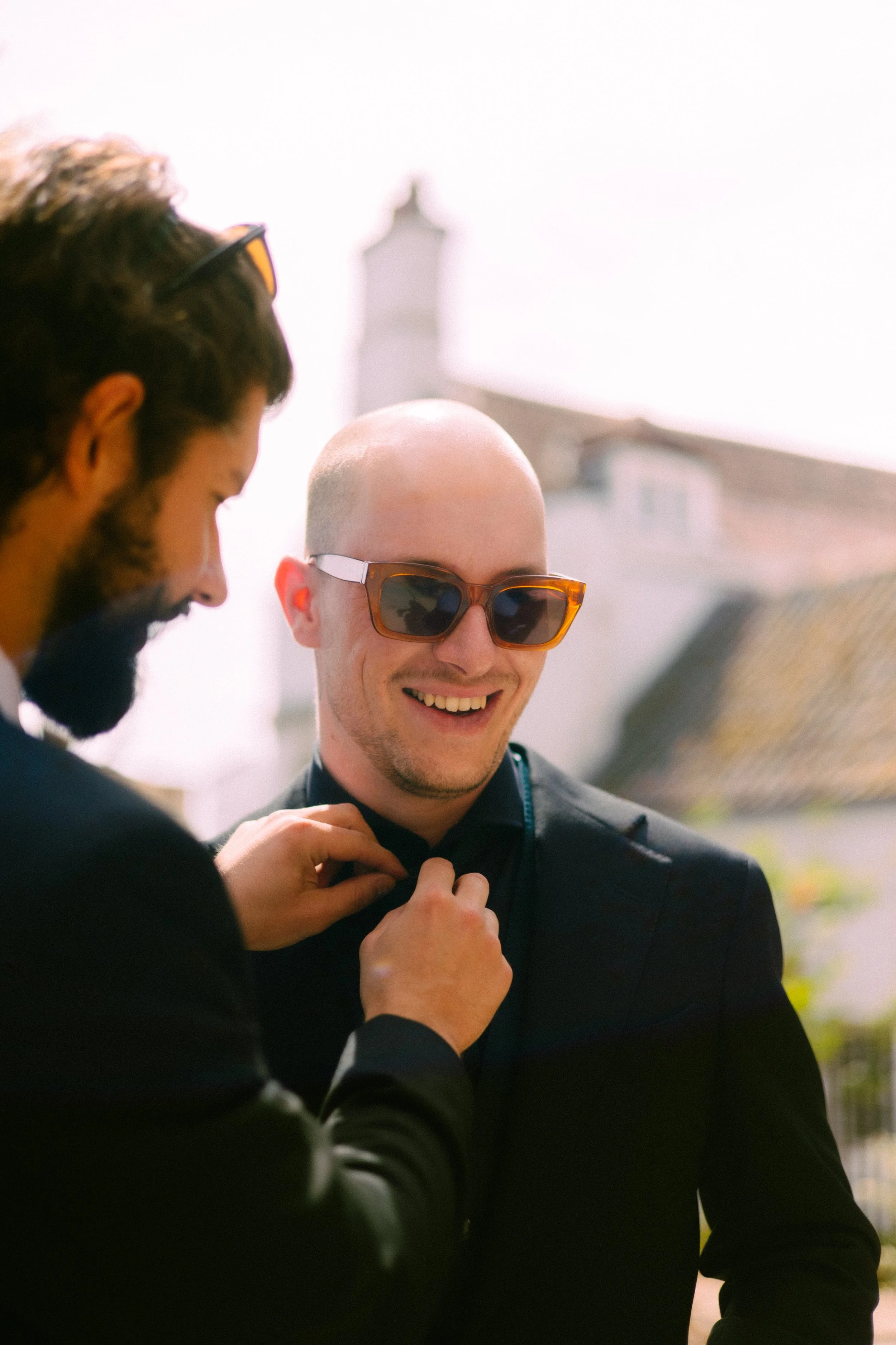 A man with a shaved head and sunglasses smiling while another person adjusts his shirt or jacket outdoors.
