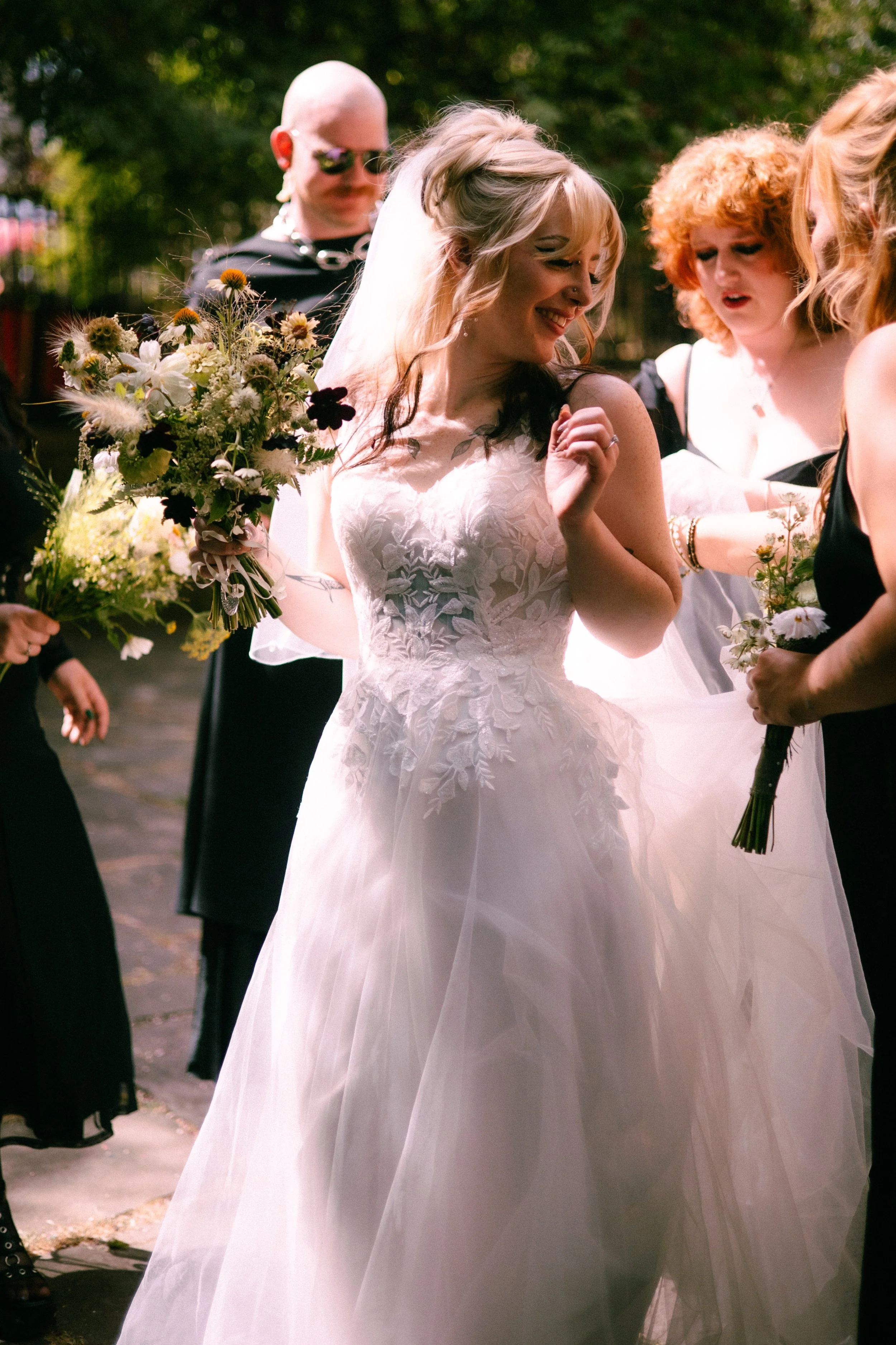 A bride in a white lace wedding dress holding a bouquet of flowers, smiling and looking down, surrounded by friends outdoors during a celebration.