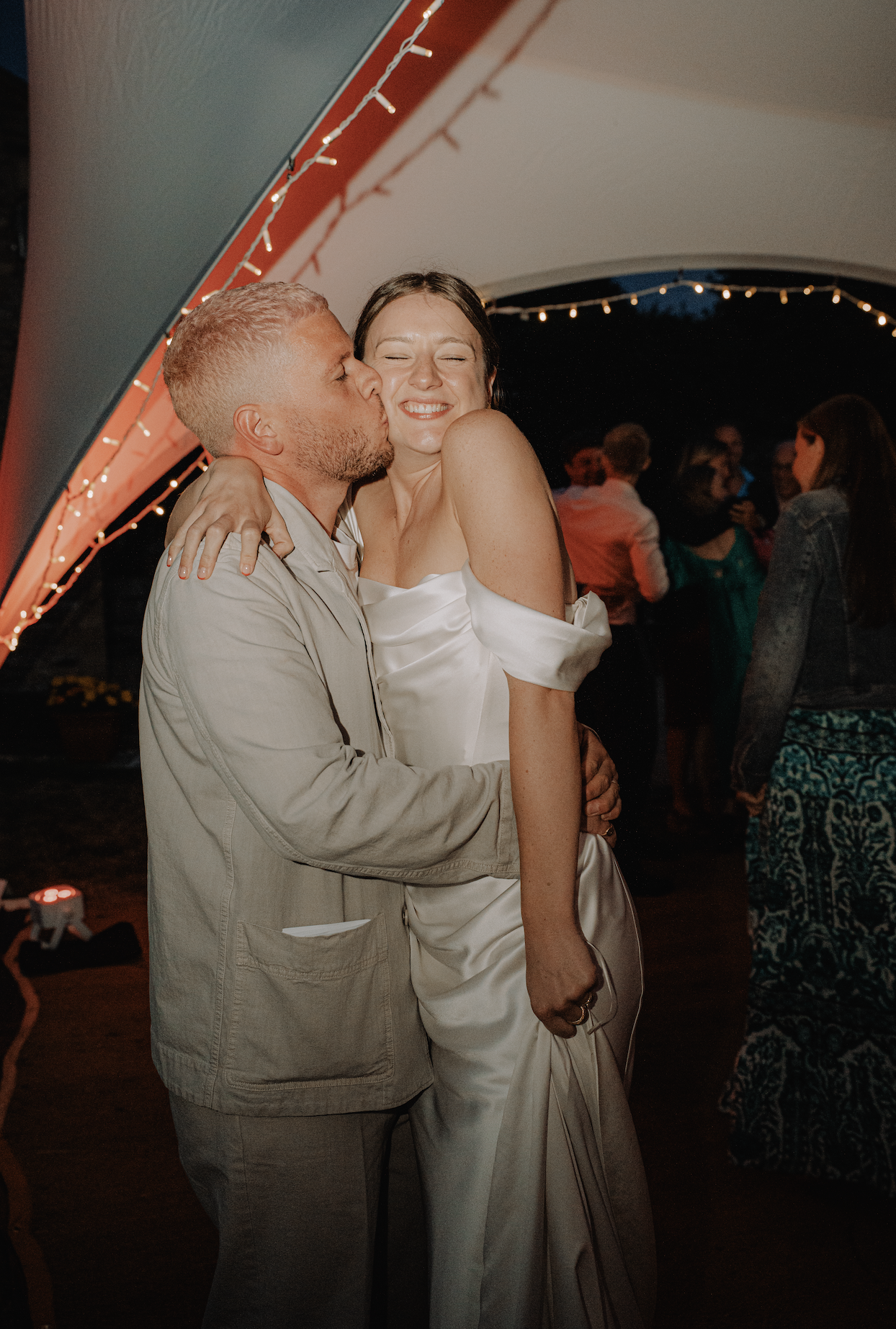 A man and woman are hugging and sharing a kiss at a celebratory event under string lights. The woman is smiling with closed eyes, dressed in a satin off-shoulder gown, and the man is in a light-colored jacket.
