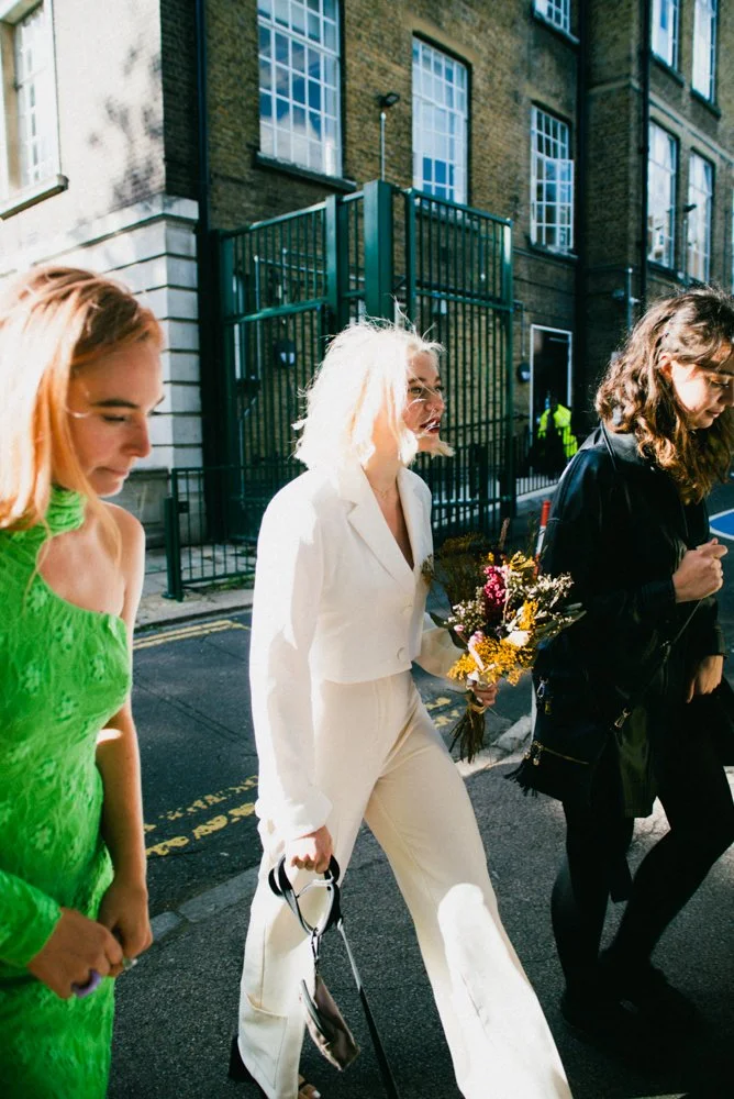 Three women walking on a street with buildings in the background. The woman in the center has platinum blonde hair, wears an all-white outfit, and carries a bouquet of flowers.