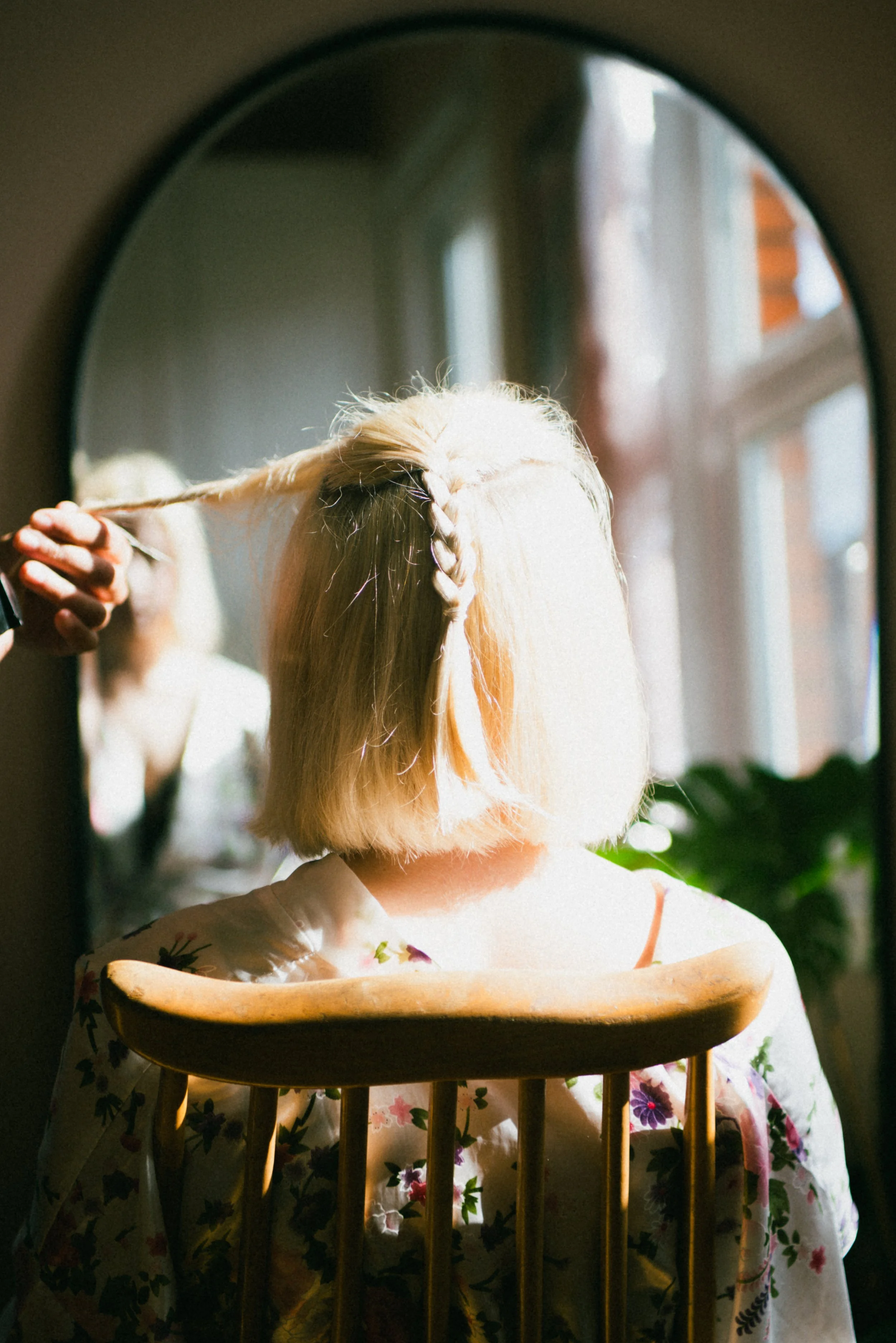 A person sitting in front of a mirror, having their hair styled. The person has short, blond hair and is wearing a floral kimono. Natural sunlight streams through the window, illuminating the scene.