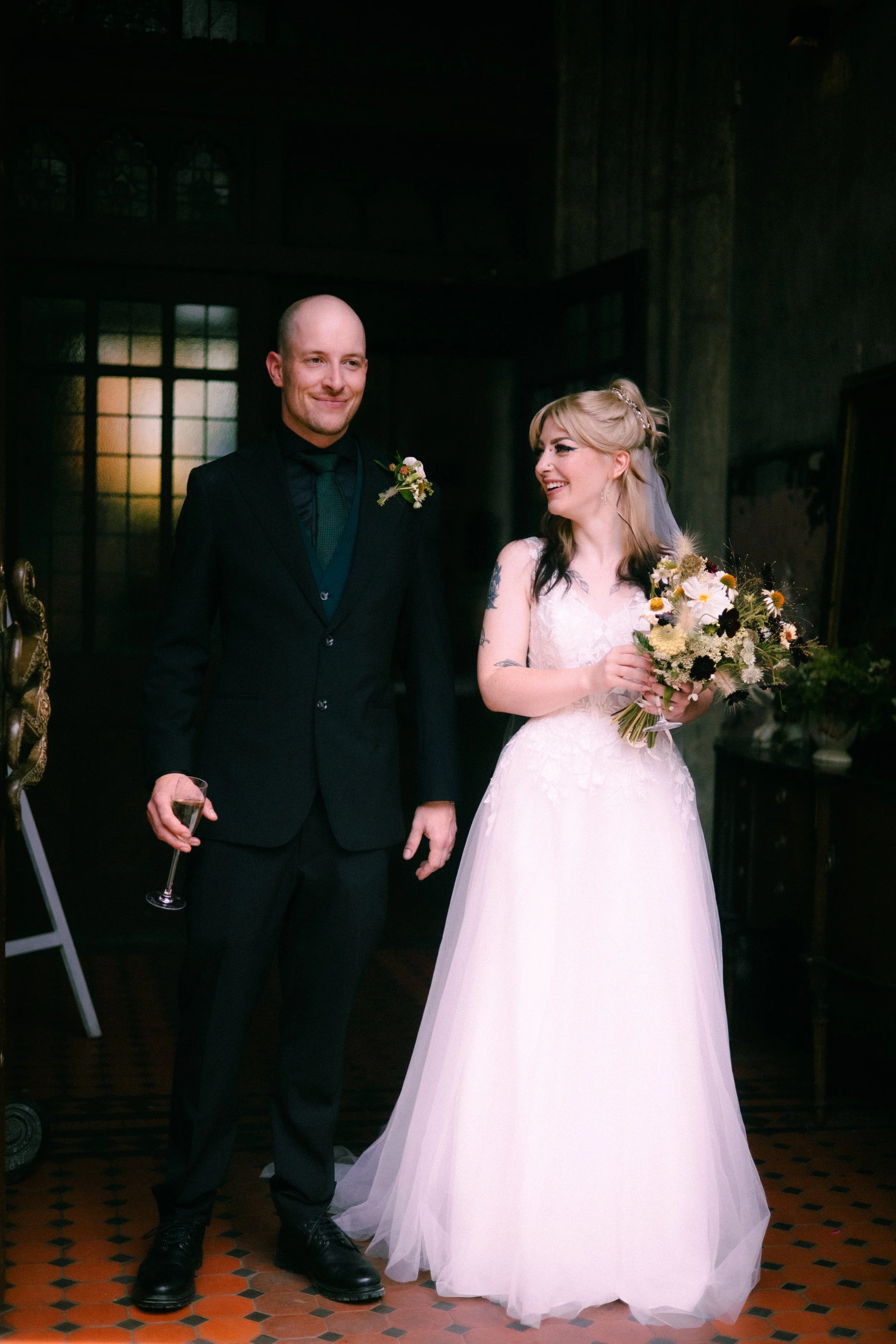 A man in a black suit and a woman in a white wedding dress standing together indoors, holding a bouquet of flowers, with the man holding a glass of champagne.