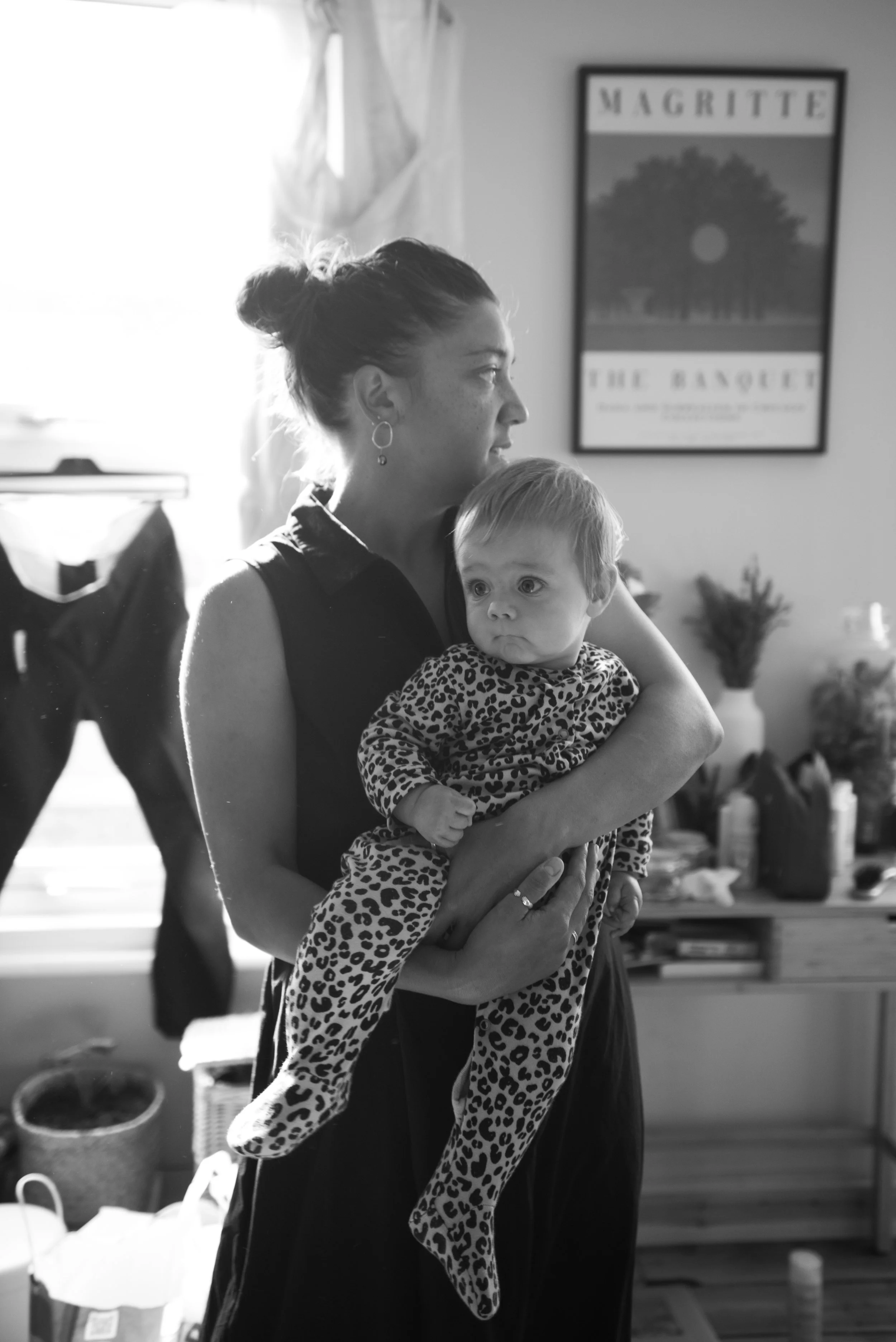 A woman holding a young girl in her arms indoors, with a window and a picture frame on the wall in the background.