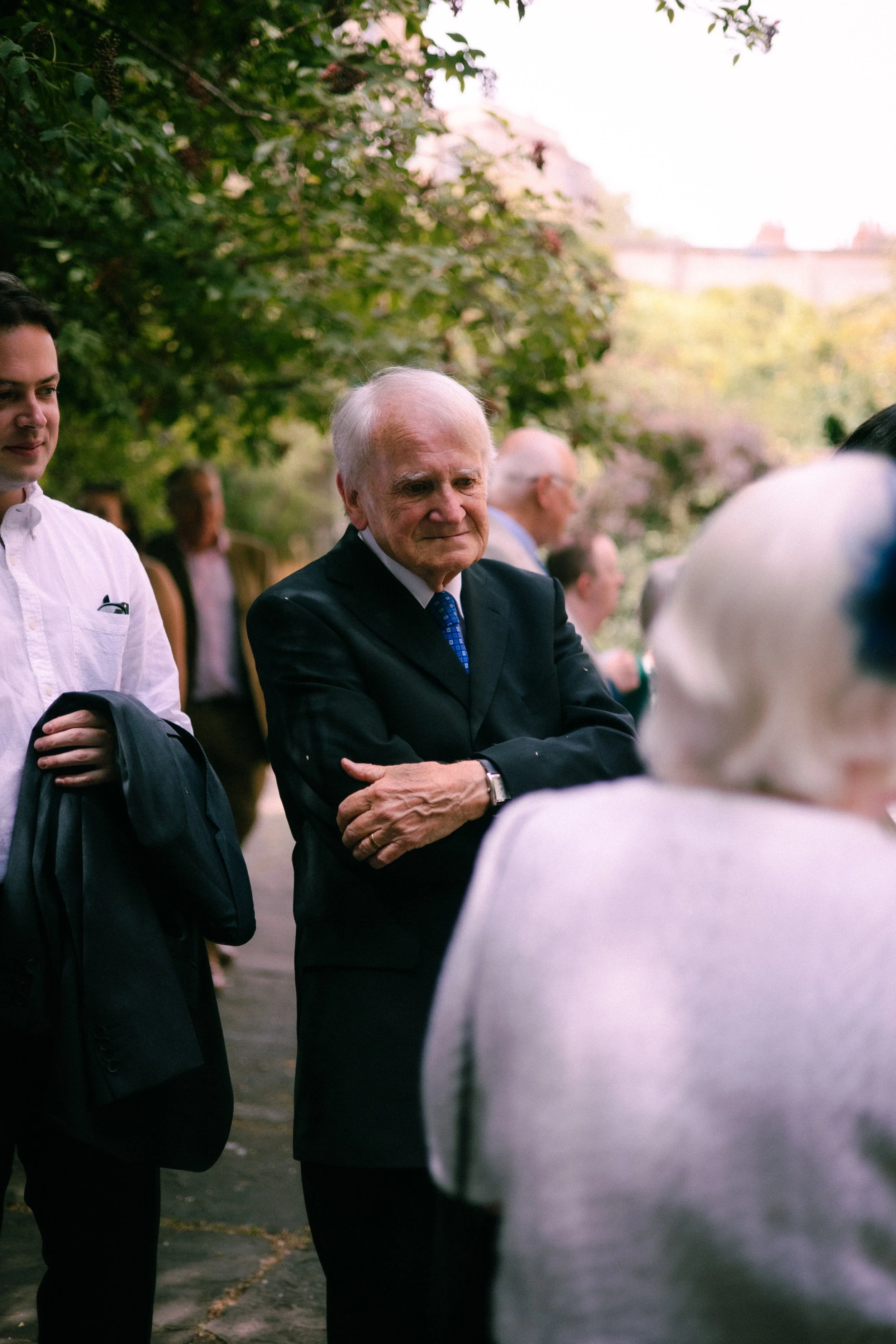 Older man in a suit talking to a woman in a white sweater outdoors, surrounded by other people under trees with pink blossoms