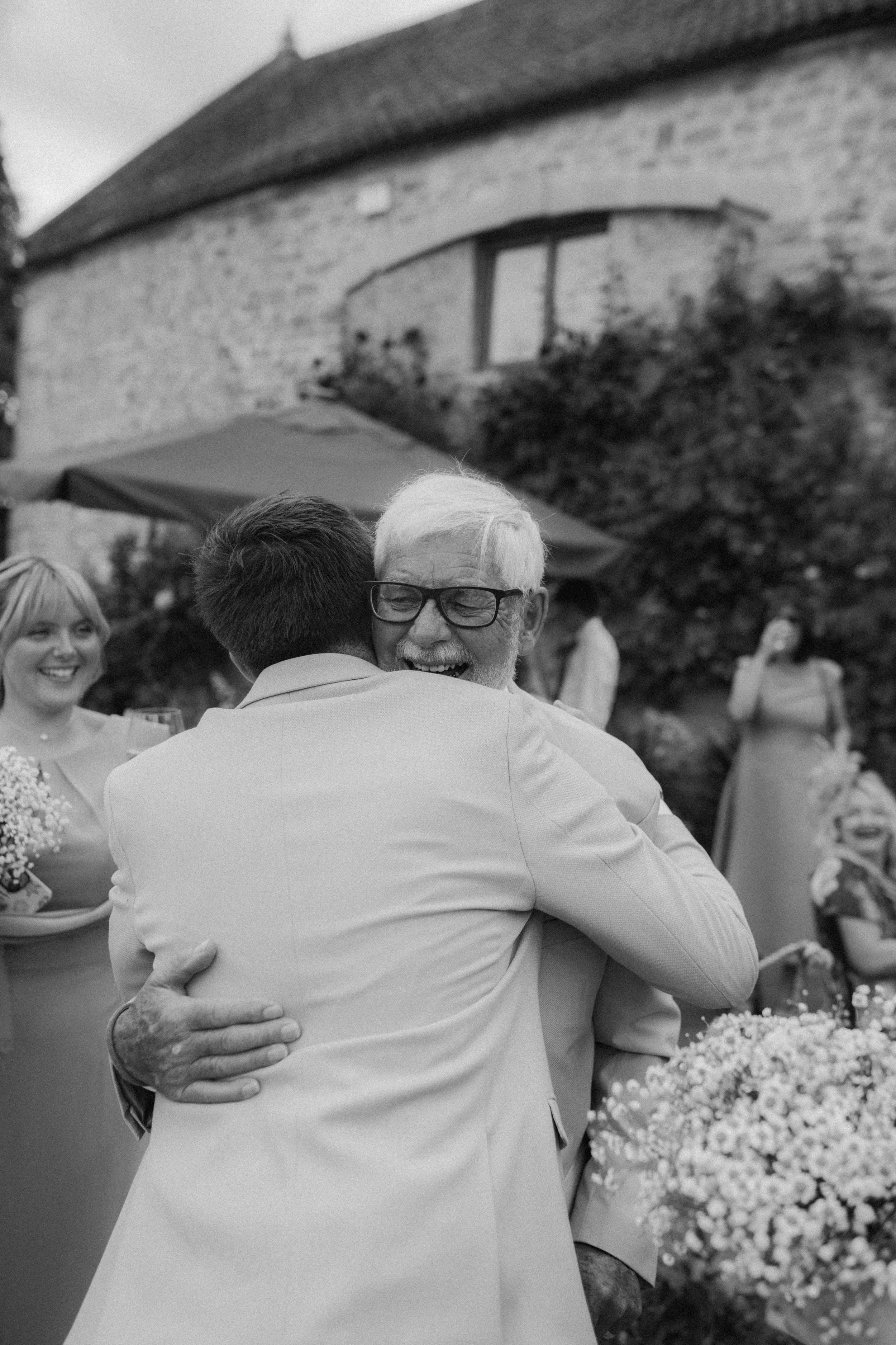 Two men hugging at an outdoor celebration, with smiling women and a building in the background.