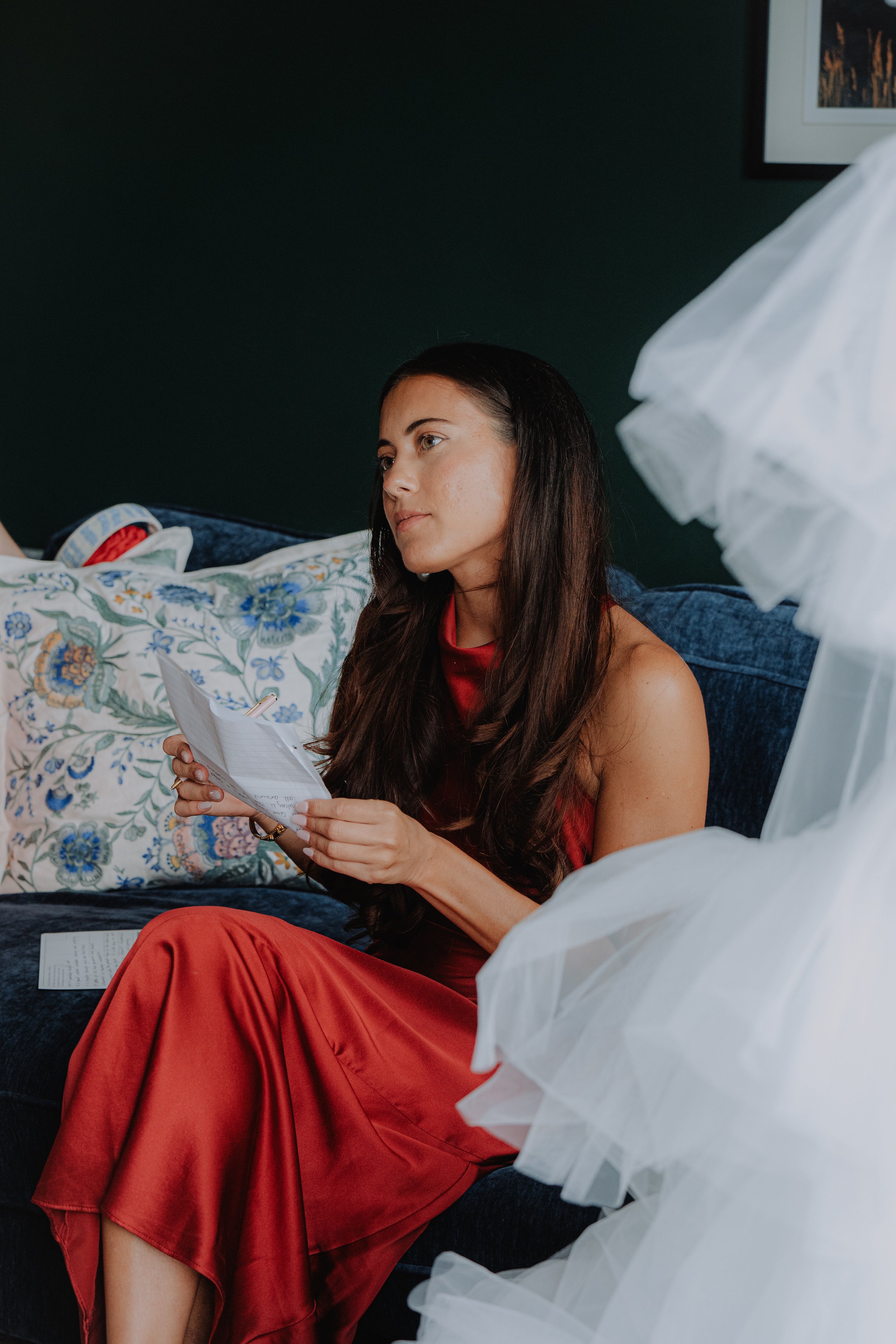 Woman in red dress sitting on a blue couch, reading a piece of paper, with floral pillow behind her and a dark green wall in the background.