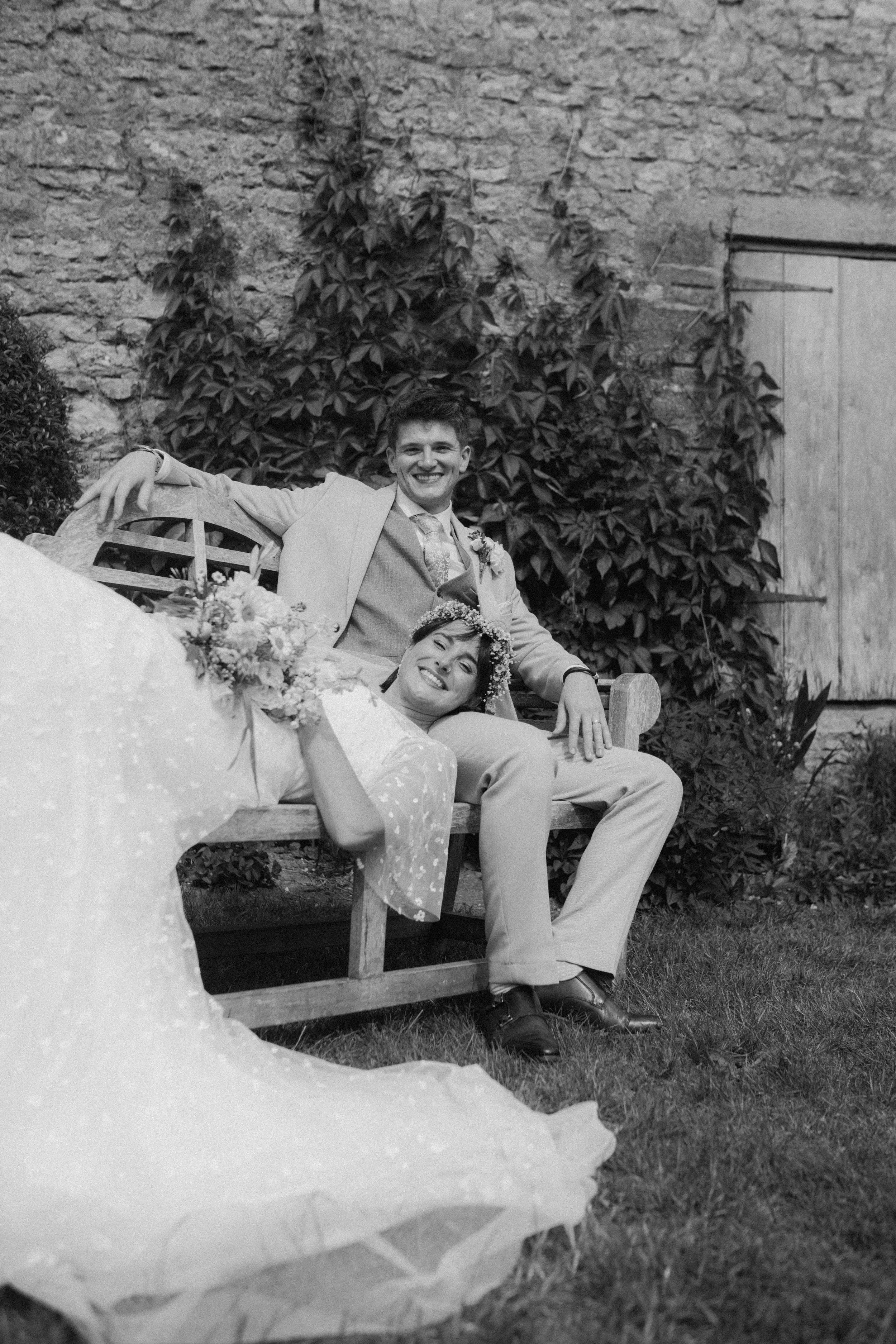 A black-and-white photo of a smiling bride and groom sitting on a wooden bench outdoors. The bride is lying down with a bouquet, wearing a floral headband and a light, patterned dress. The groom is sitting beside her, wearing a light-colored suit, lo