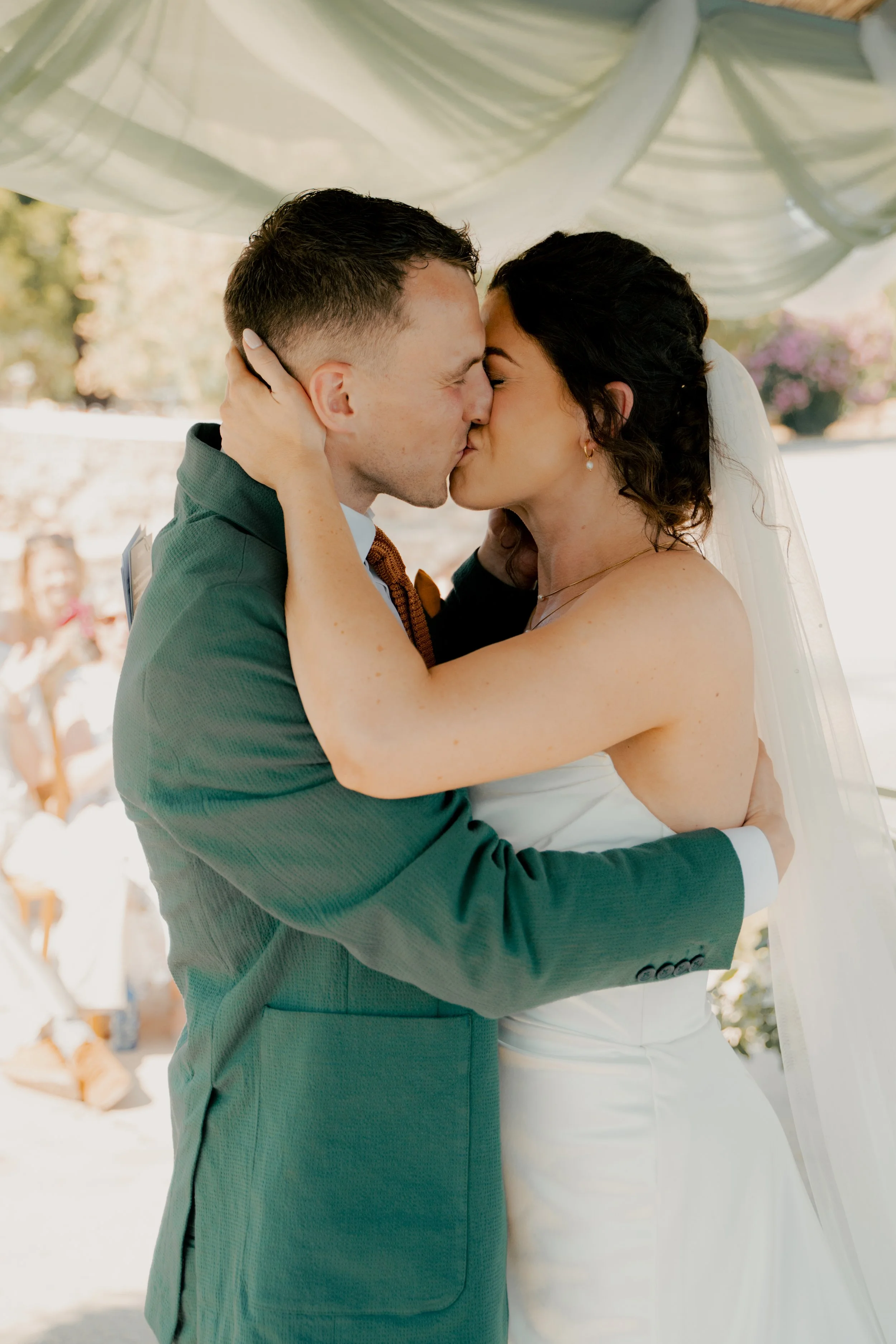A bride and groom share a kiss during their outdoor wedding ceremony, with wedding guests in the background.