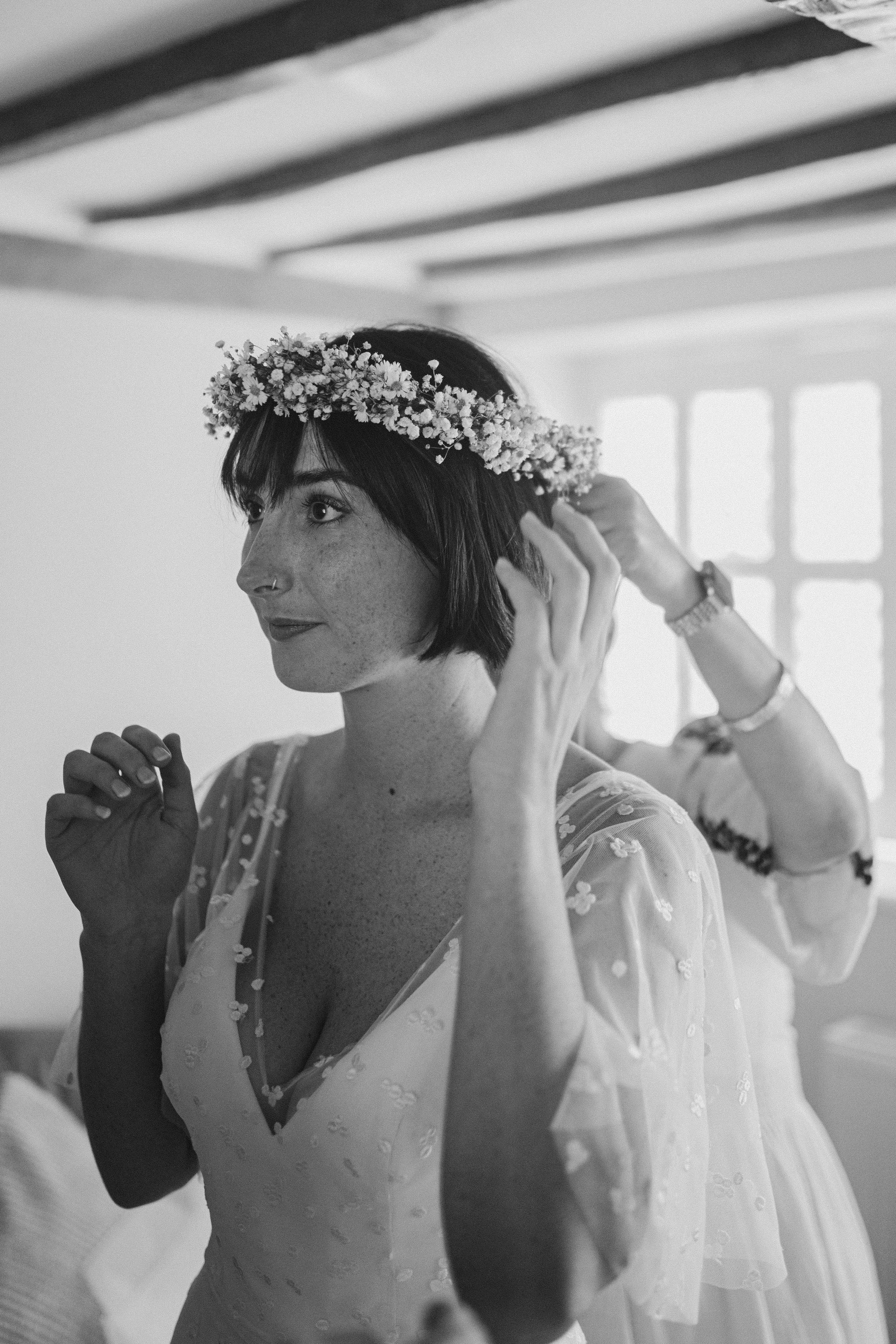 Portrait of a woman with short dark hair wearing a floral crown and a light dress, indoors near a window.