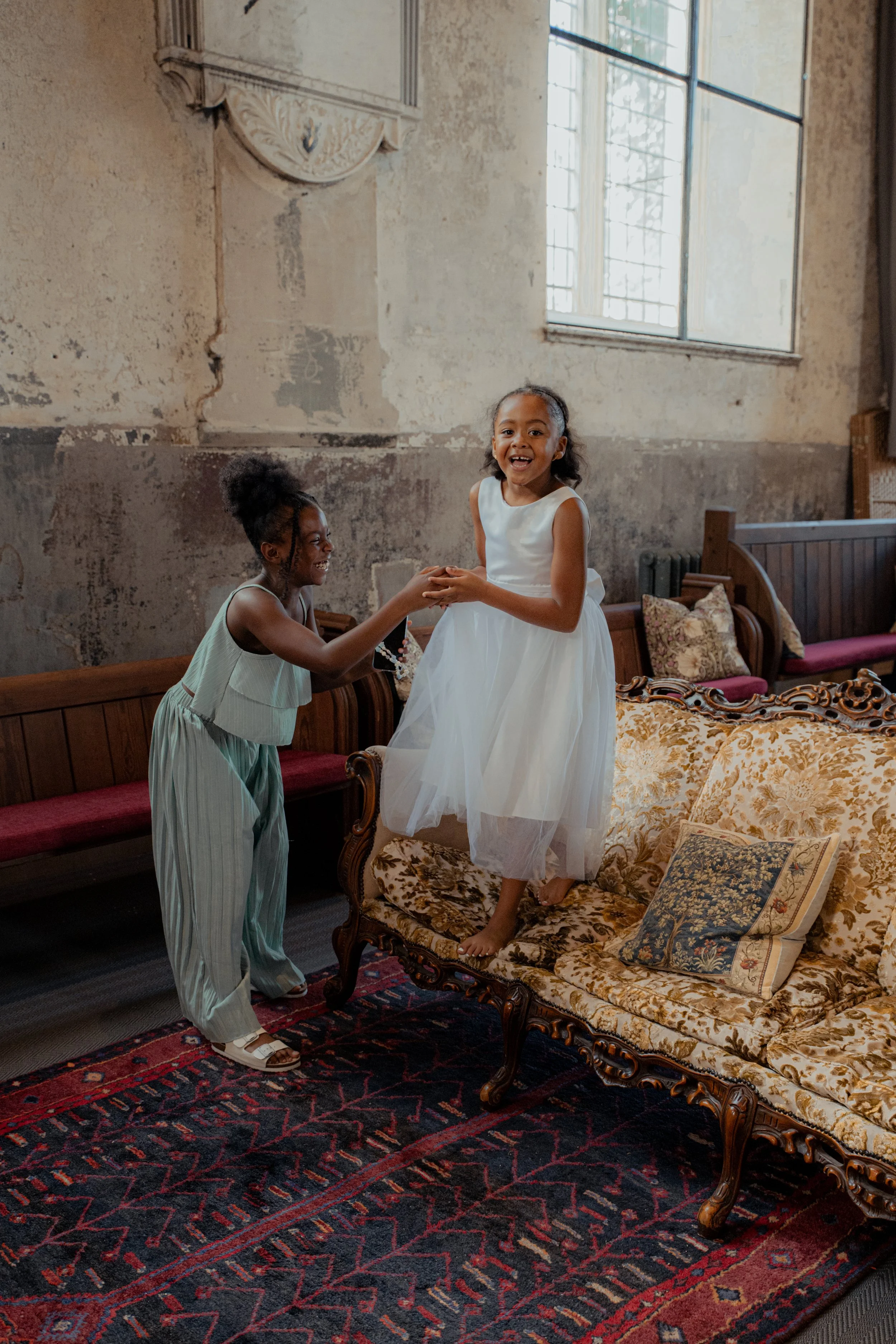Two young girls, one standing on a vintage floral upholstered sofa and the other on the floor, exchanging smiles and hand gestures in a room with distressed walls, a large window, and vintage furniture.