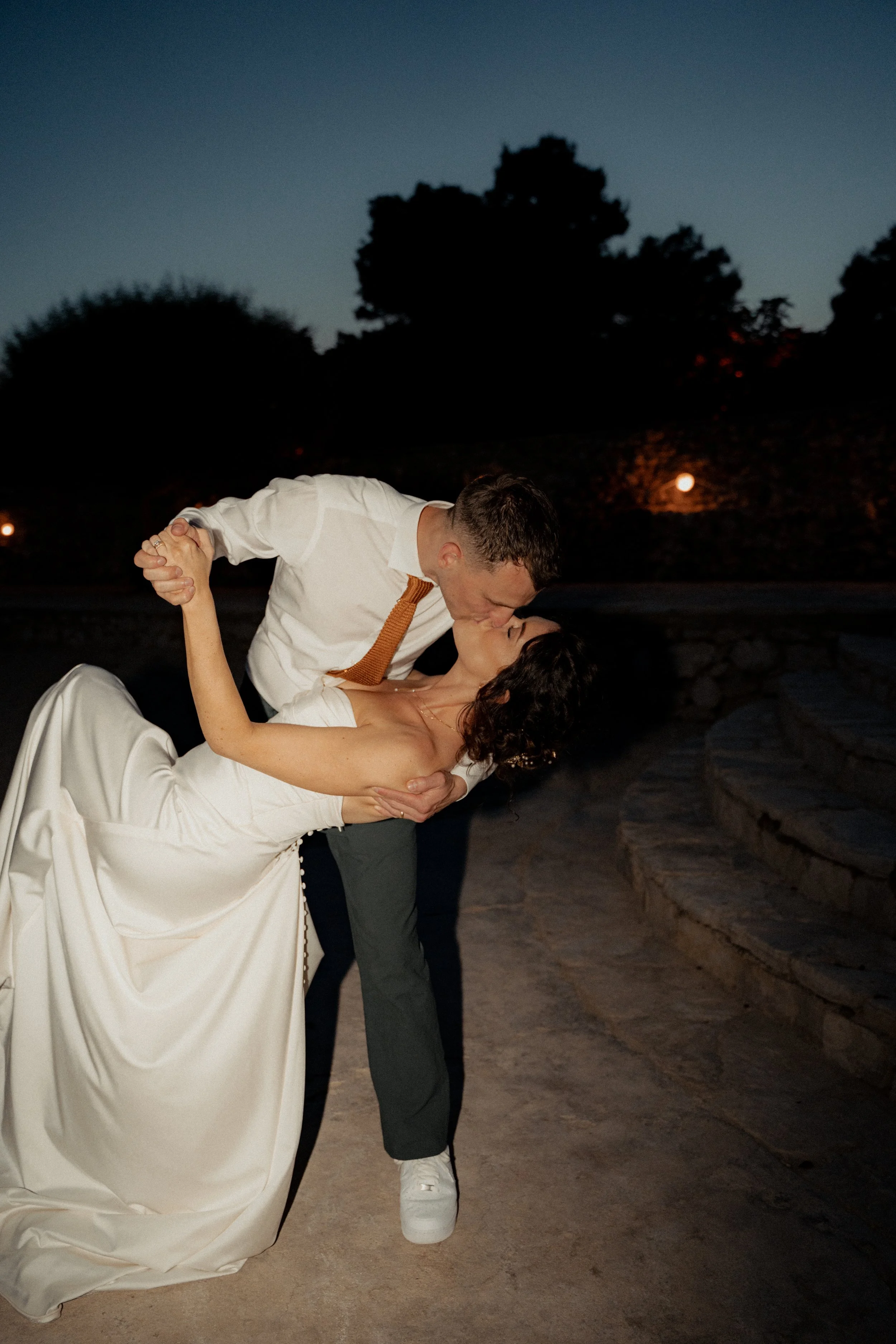 A couple shares a romantic kiss during their wedding photoshoot at dusk, with the man dipping the woman in a white wedding dress.