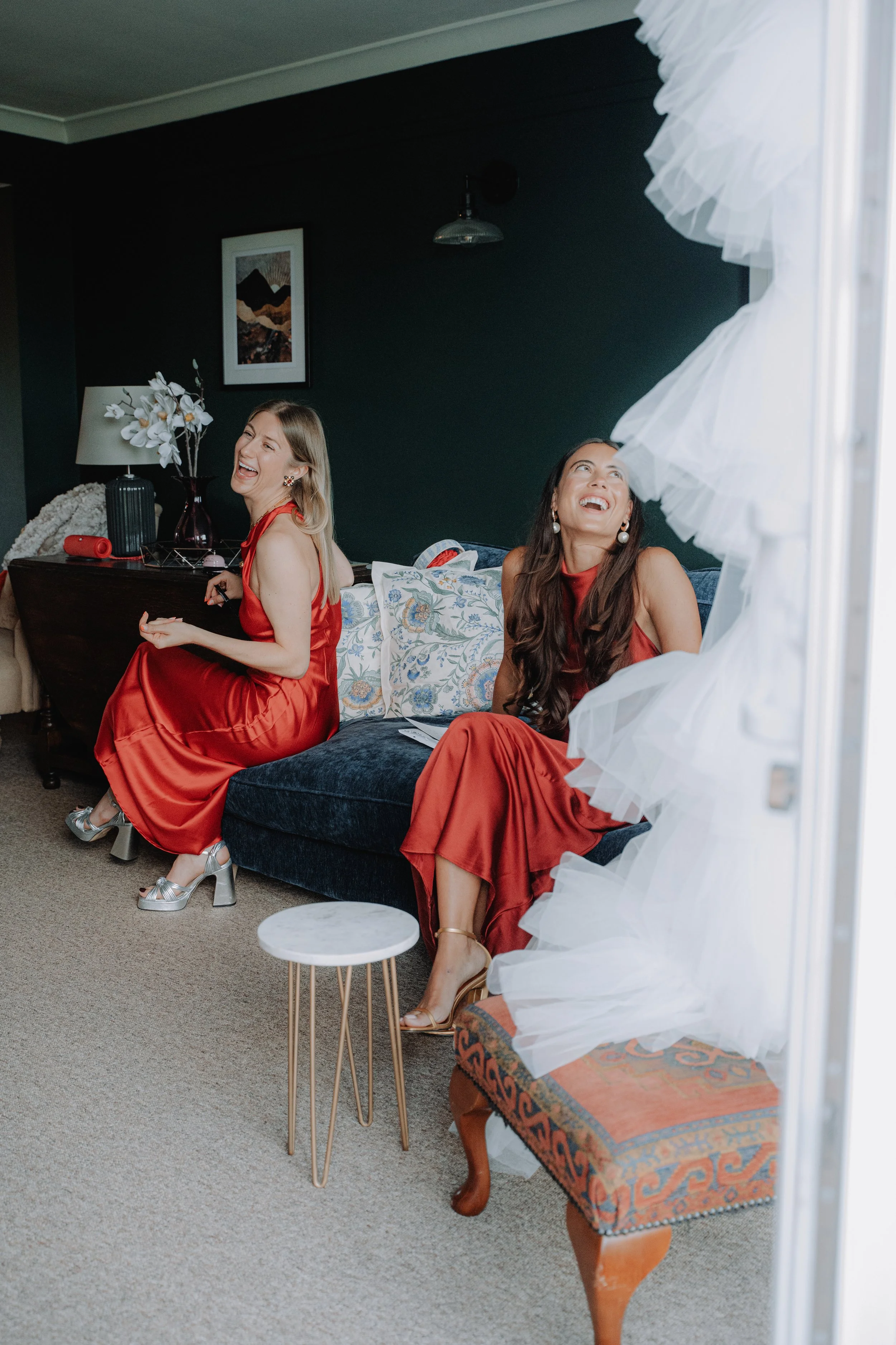 Two women in red dresses laughing and sitting on a dark blue couch in a stylish living room with dark walls, white curtains, and decorative pillows.