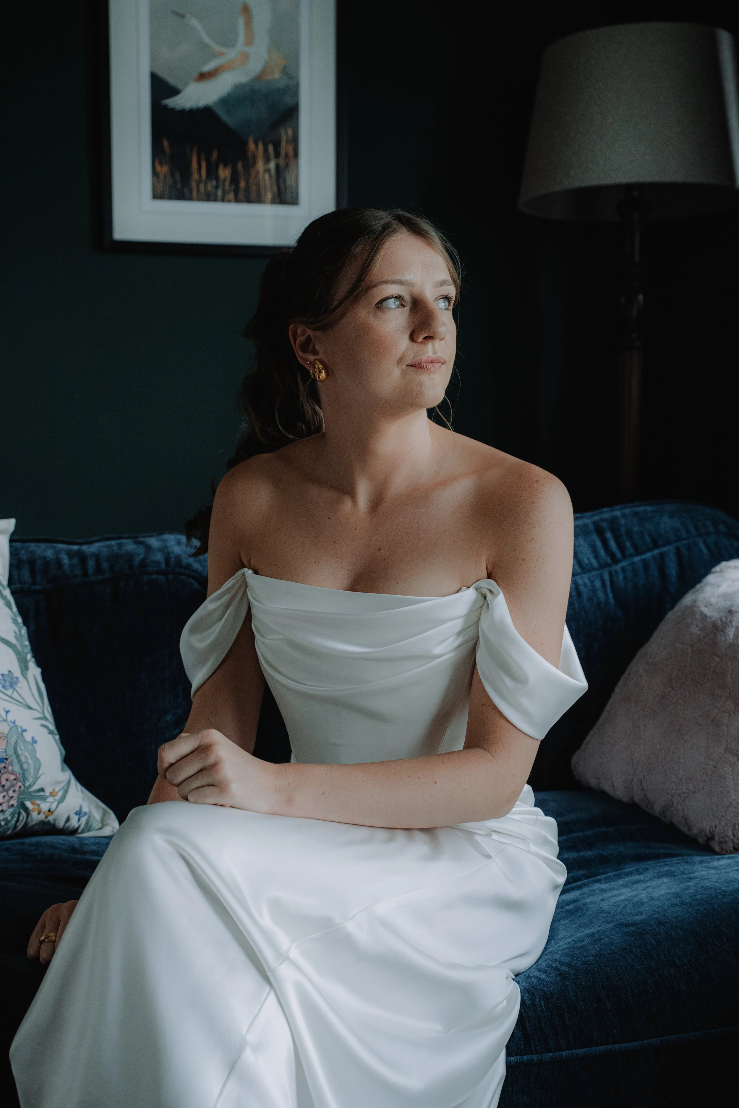 A woman in a white off-the-shoulder dress sitting on a dark blue couch, looking to her right, with a painting of a bird flying over water hanging on a dark wall behind her.