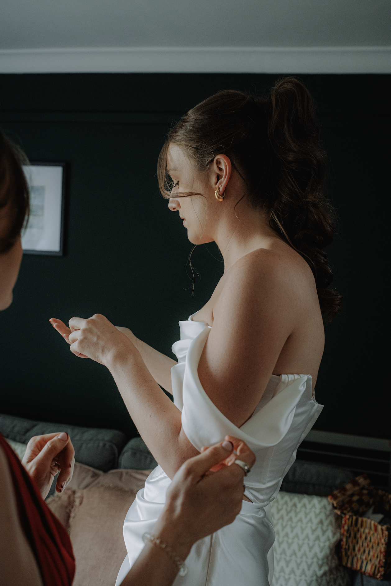 A woman in an off-shoulder satin dress is getting ready with assistance from another person, possibly her wedding or special occasion prep, inside a room with dark walls and plush pillows.