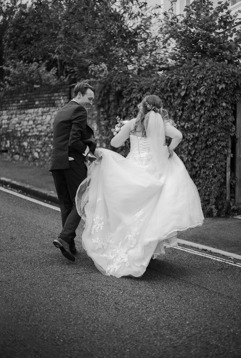 A bride and groom share a joyful moment outdoors, with the bride lifting her wedding gown as the groom helps her walk. Black and white photograph.