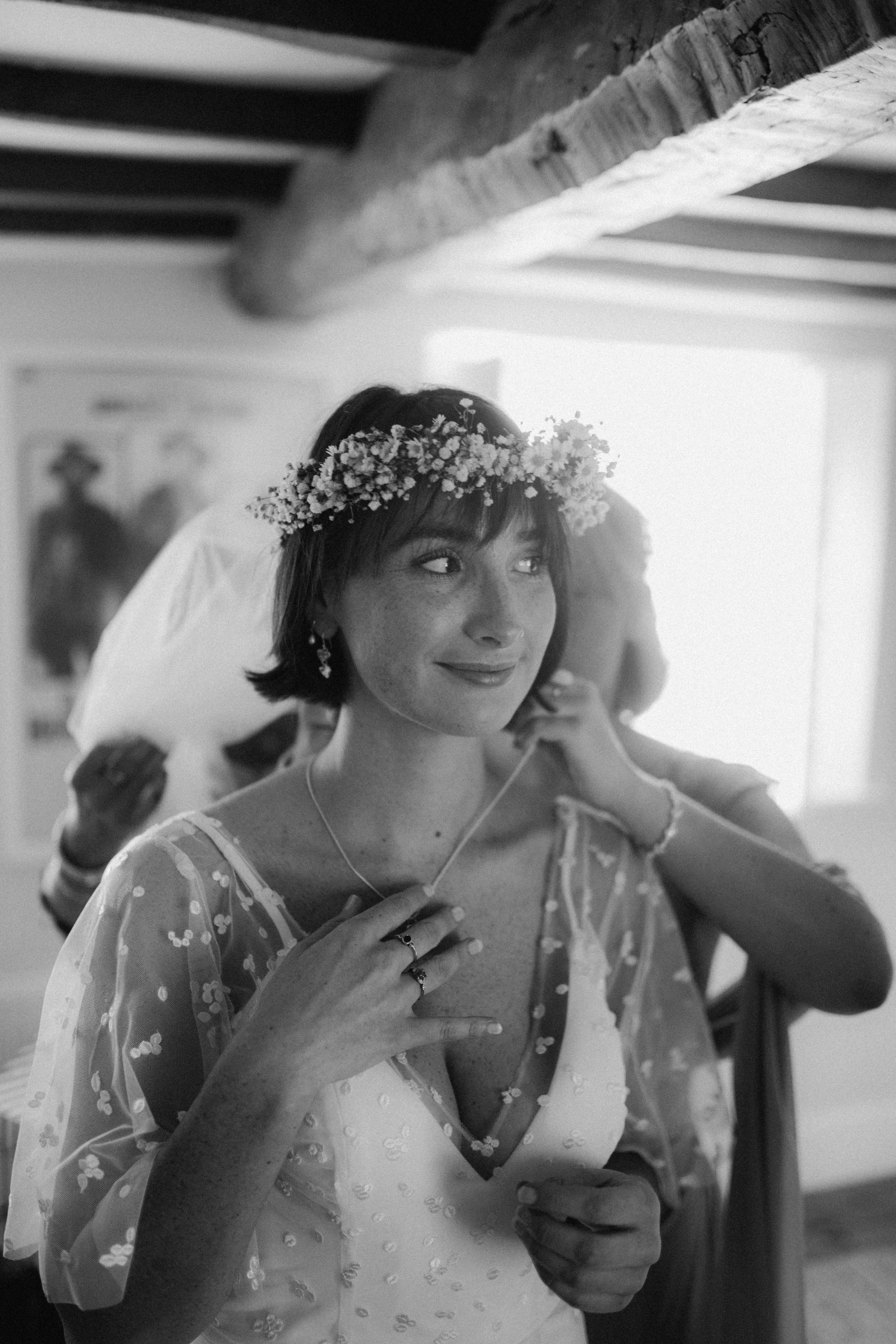 Black and white photo of a woman wearing a flower crown, bracelet, and dress with a sheer overlay, holding a necklace near her neck, with a person adjusting a veil behind her.