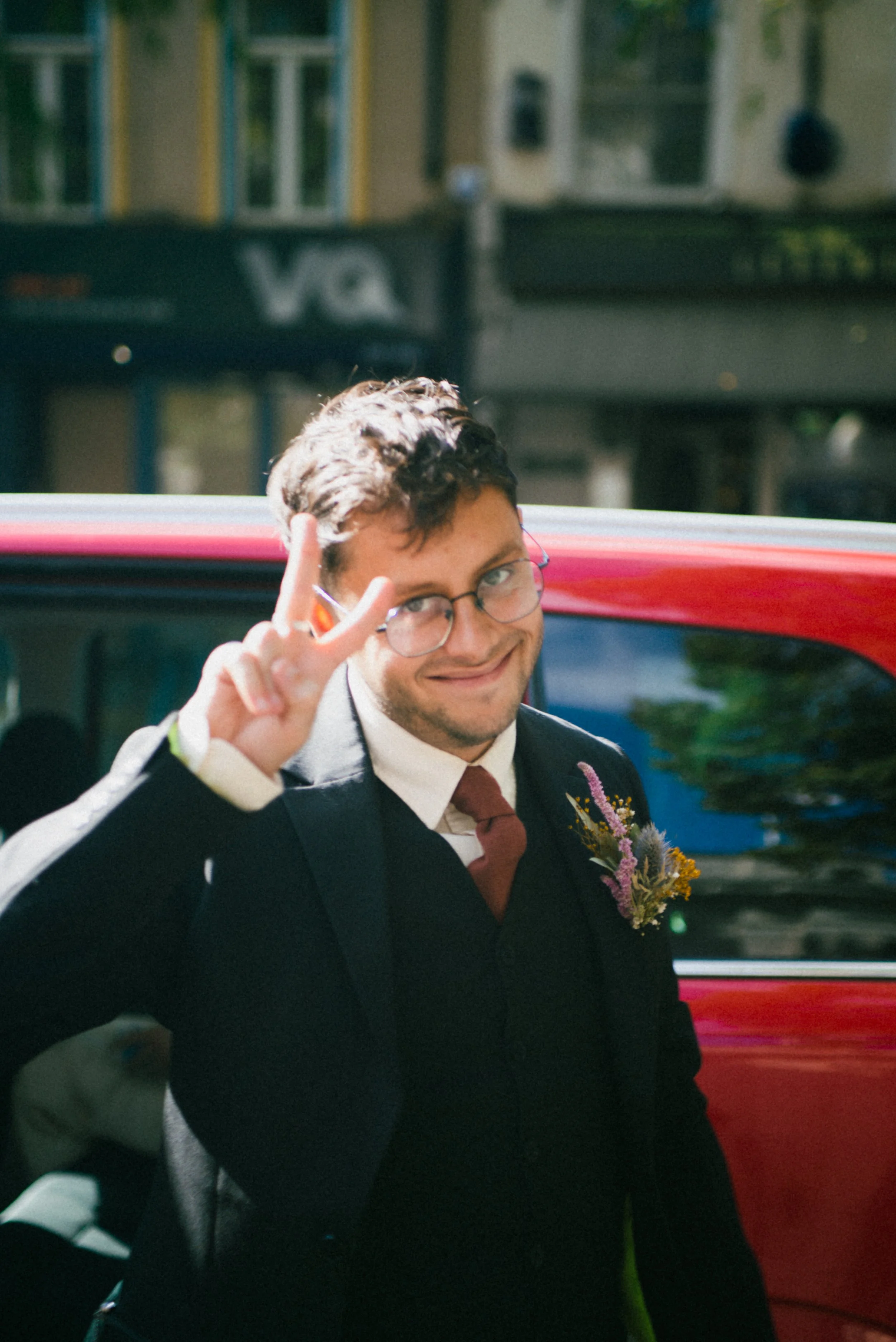 A man in a suit with glasses making a peace sign and smiling outdoors next to a red car.