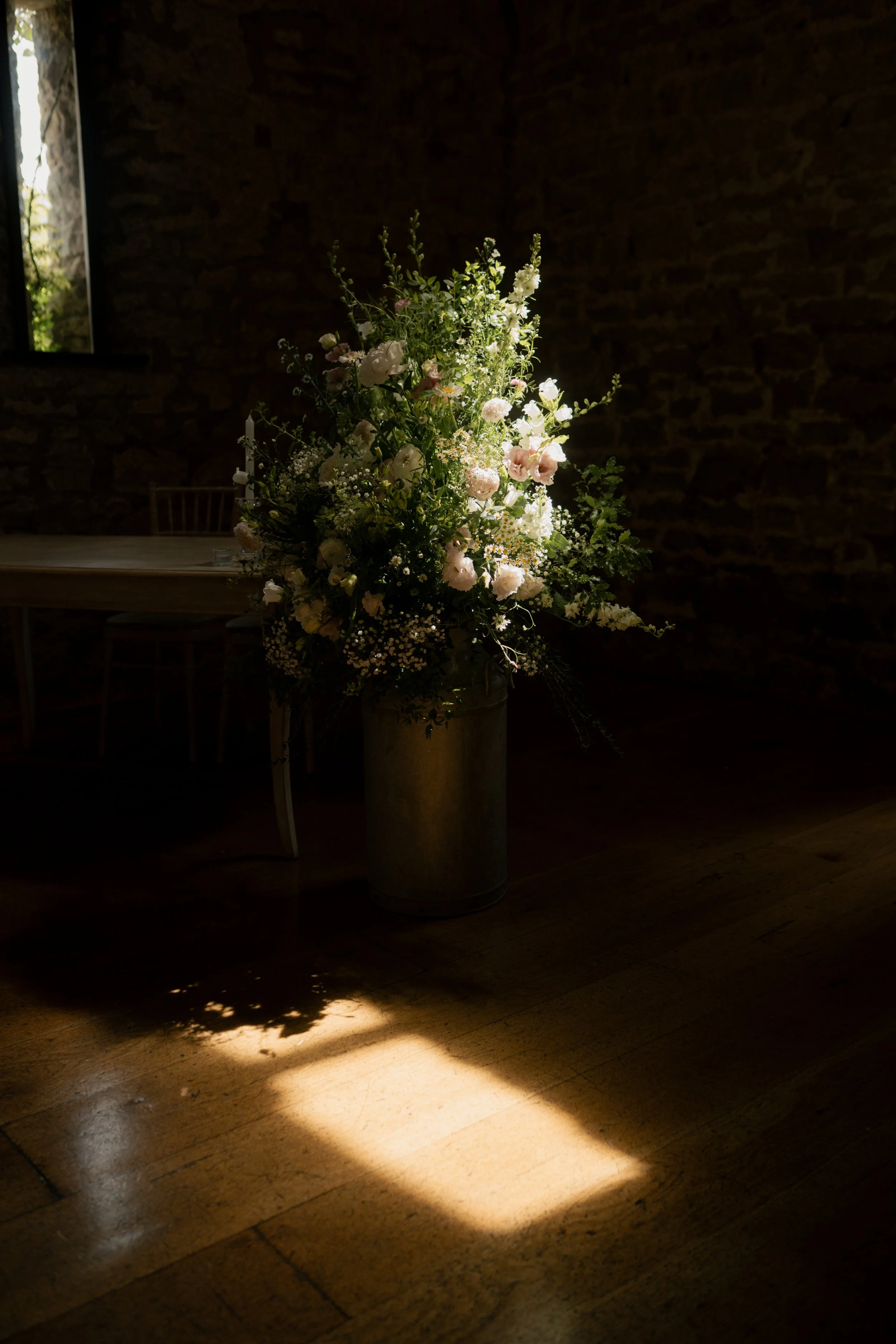 A tall floral arrangement with white, pink, and green flowers in a metal vase, illuminated by a beam of sunlight on a wooden floor, with a brick wall in the background.