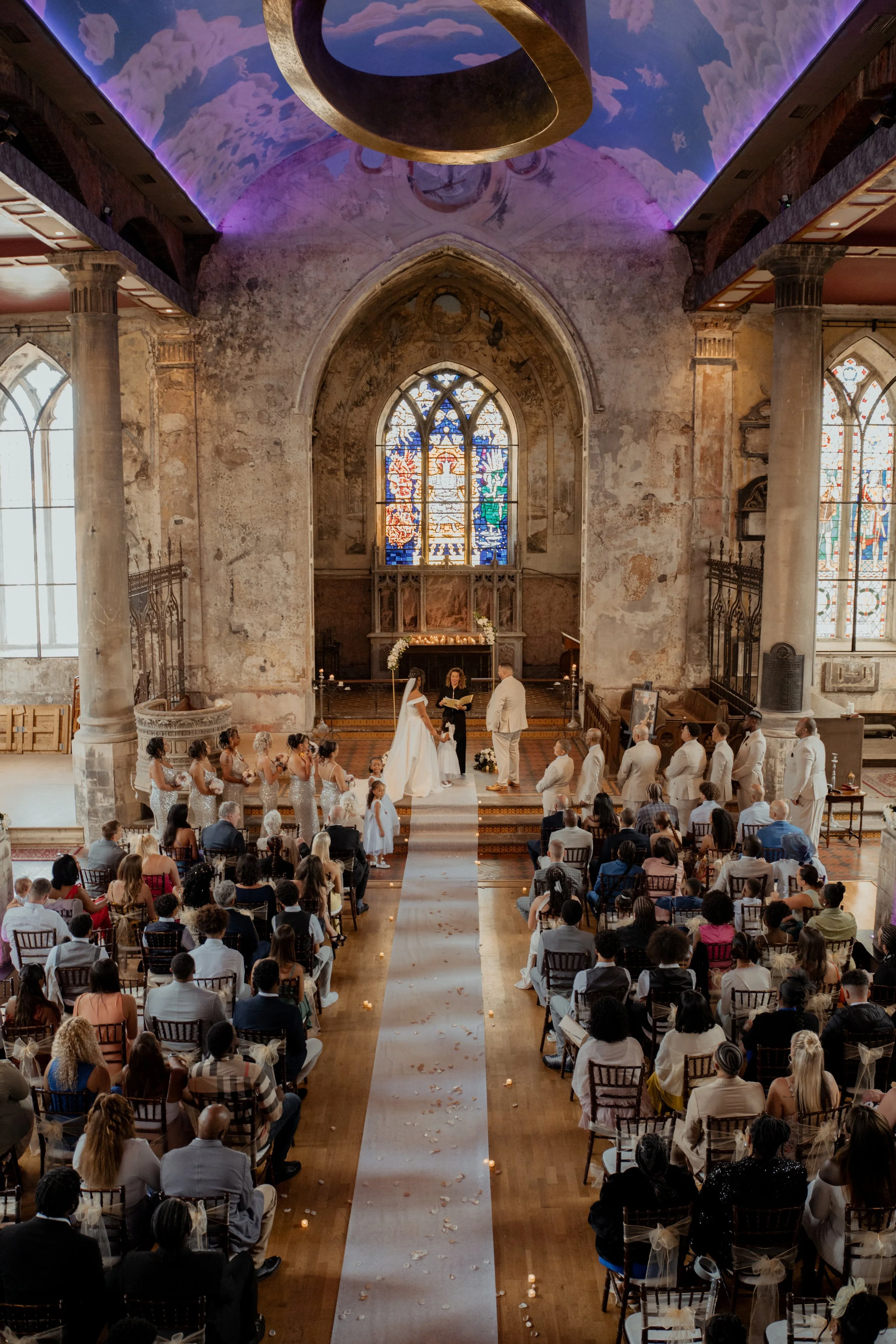 A wedding ceremony taking place inside a historic church with stained glass windows and exposed, weathered stone walls. The bride and groom are at the altar, with bridesmaids and groomsmen standing nearby. Guests are seated on wooden chairs, watching