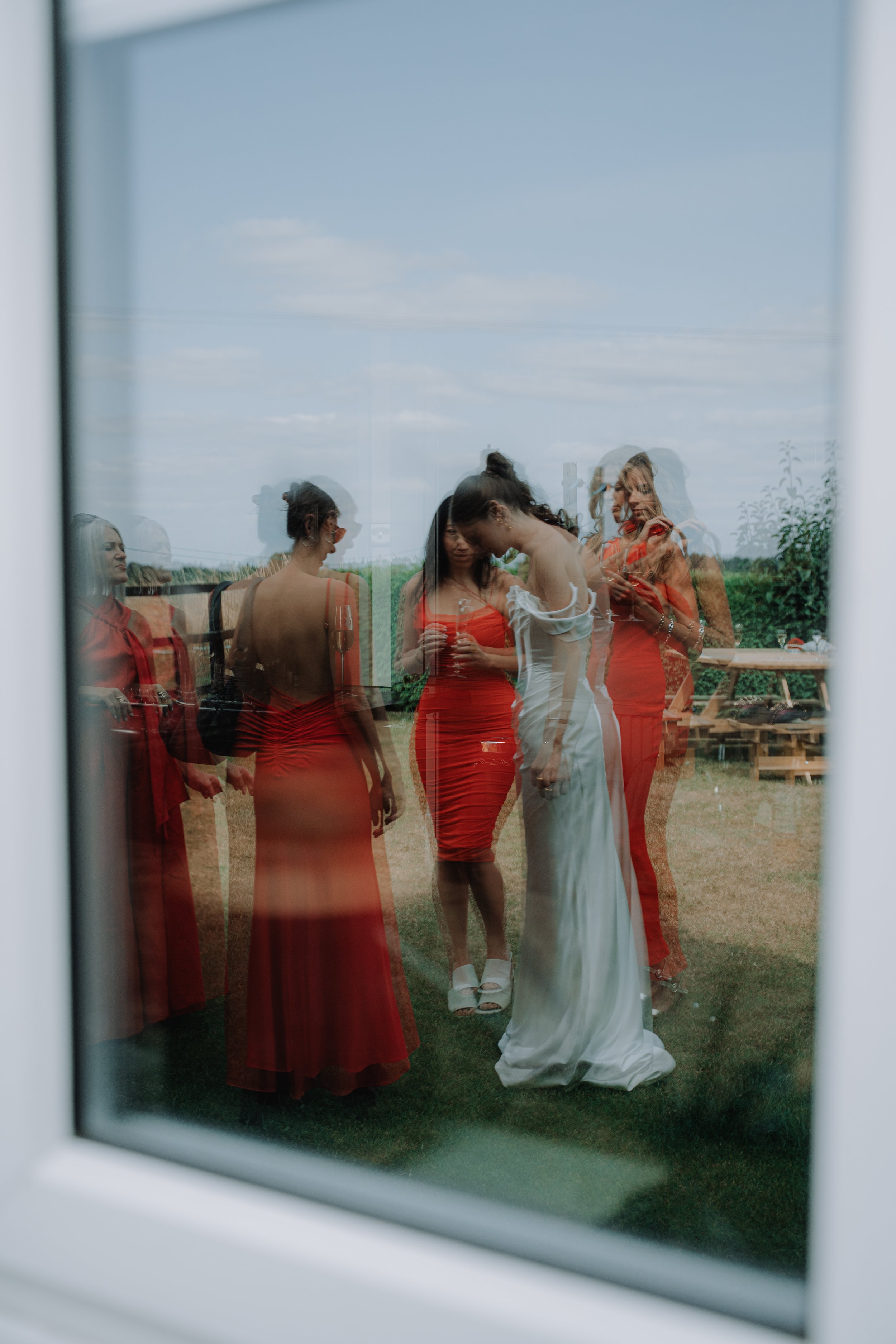 Women in red dresses and a woman in a white dress reflected in a window, outside on a grassy area with a picnic table, under a cloudy sky.