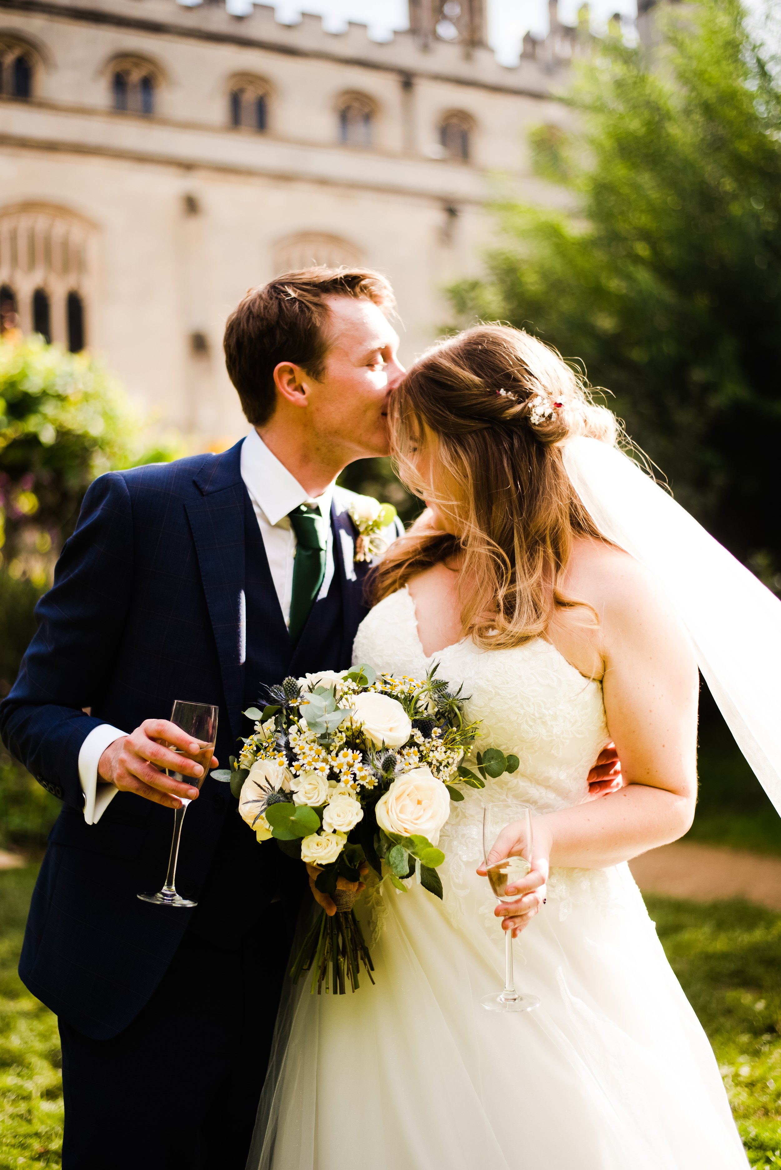 A newlywed couple sharing a kiss outdoors, with the groom in a dark suit and the bride in a white wedding gown holding a bouquet of white roses and greenery, both holding champagne flutes.
