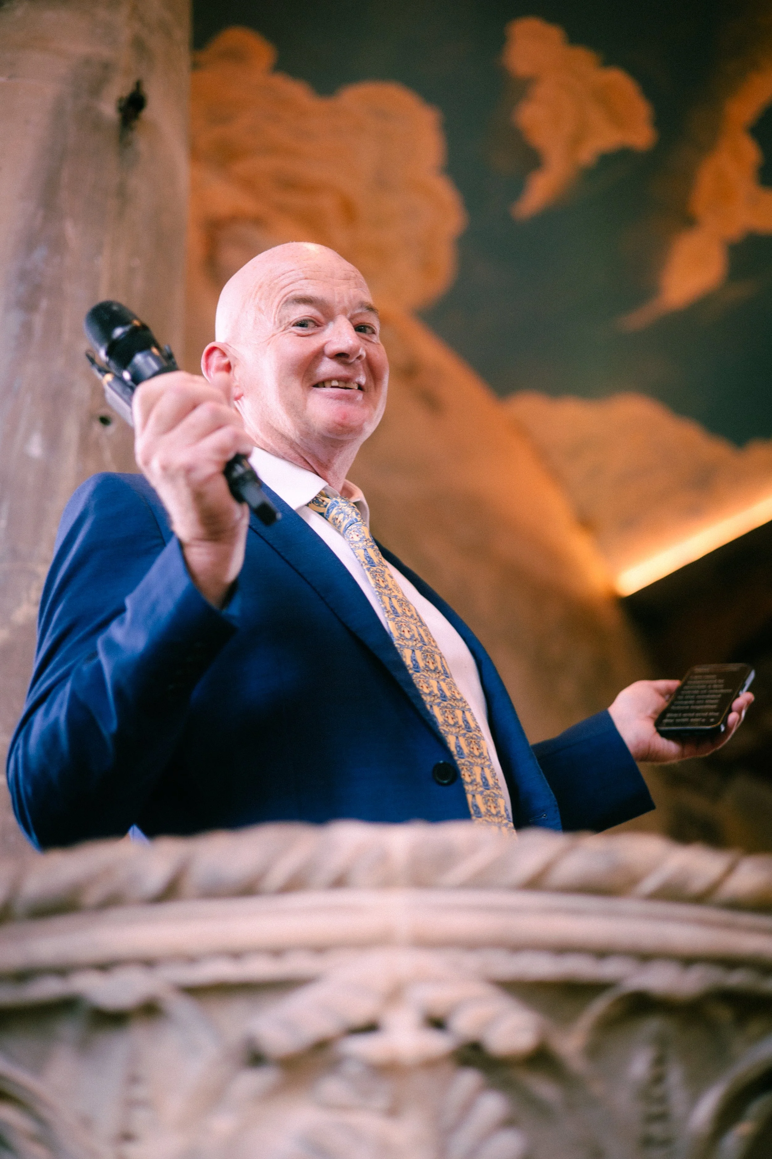 A smiling man in a navy blue suit, white shirt, and patterned tie, holding a microphone in one hand and a smartphone in the other, standing indoors with a large, orange abstract painting behind him.