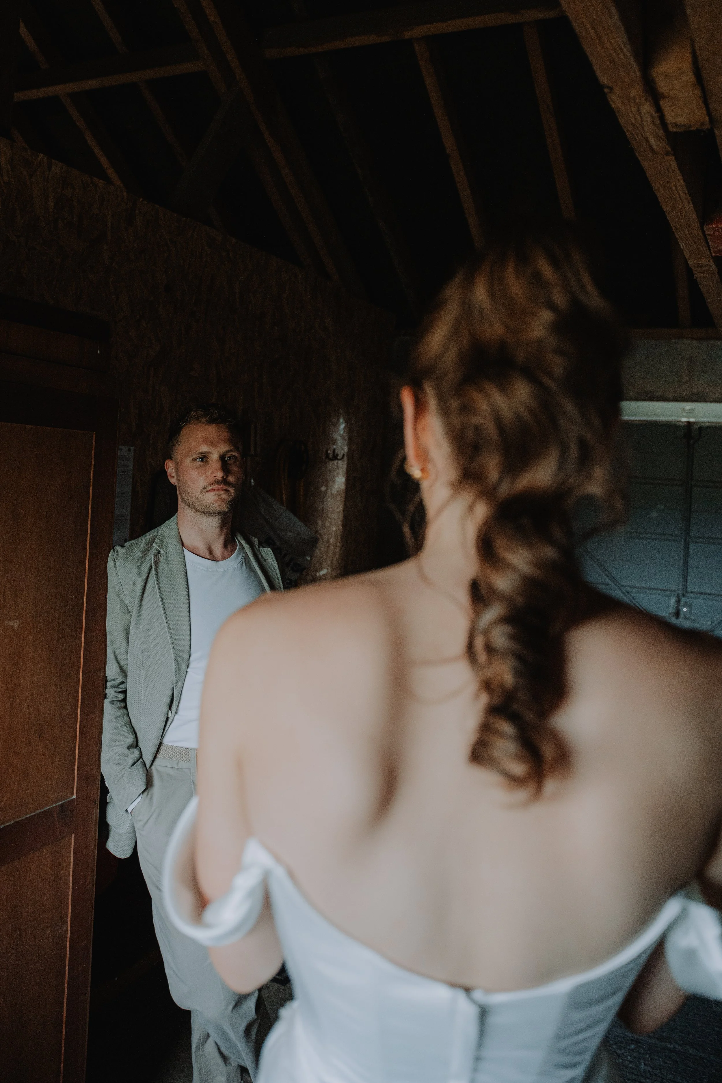 A woman in a white dress with off-shoulder sleeves faces a man in a beige suit, standing in a wooden room with an exposed ceiling. They appear to be looking at each other intently.