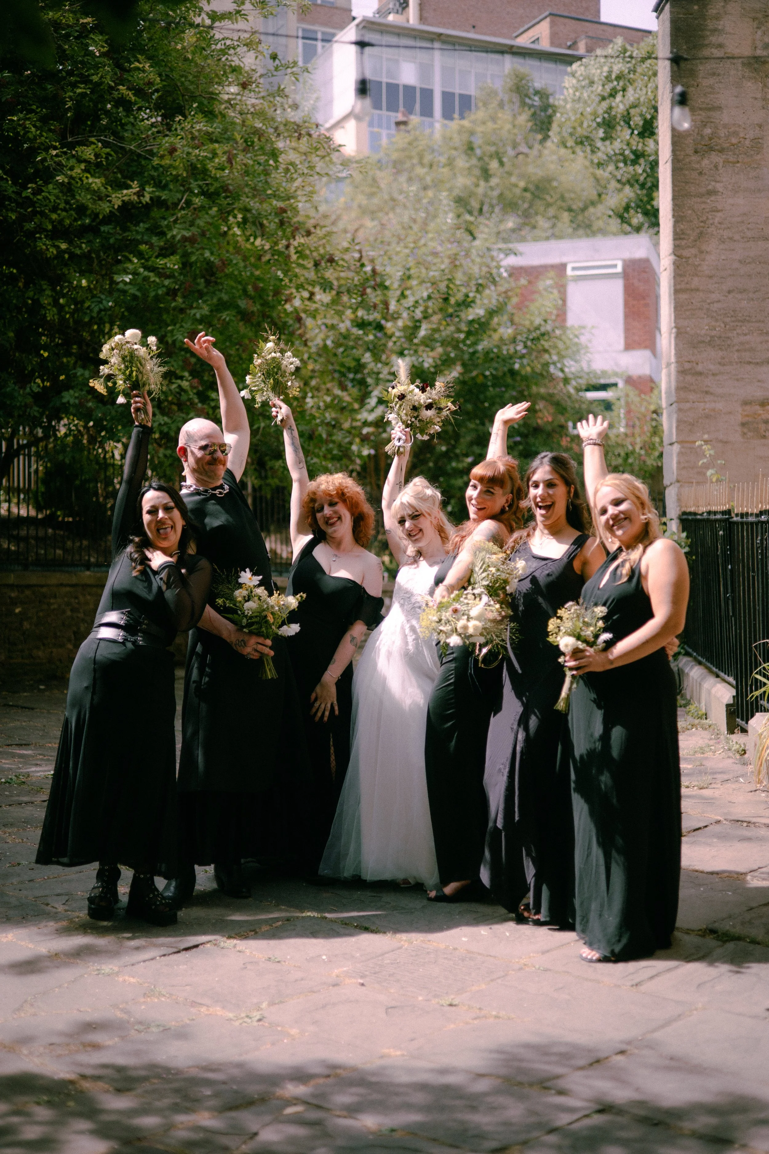 Group of people dressed in black and white, smiling and celebrating outdoors on a sunny day, holding bouquets and raising their hands.