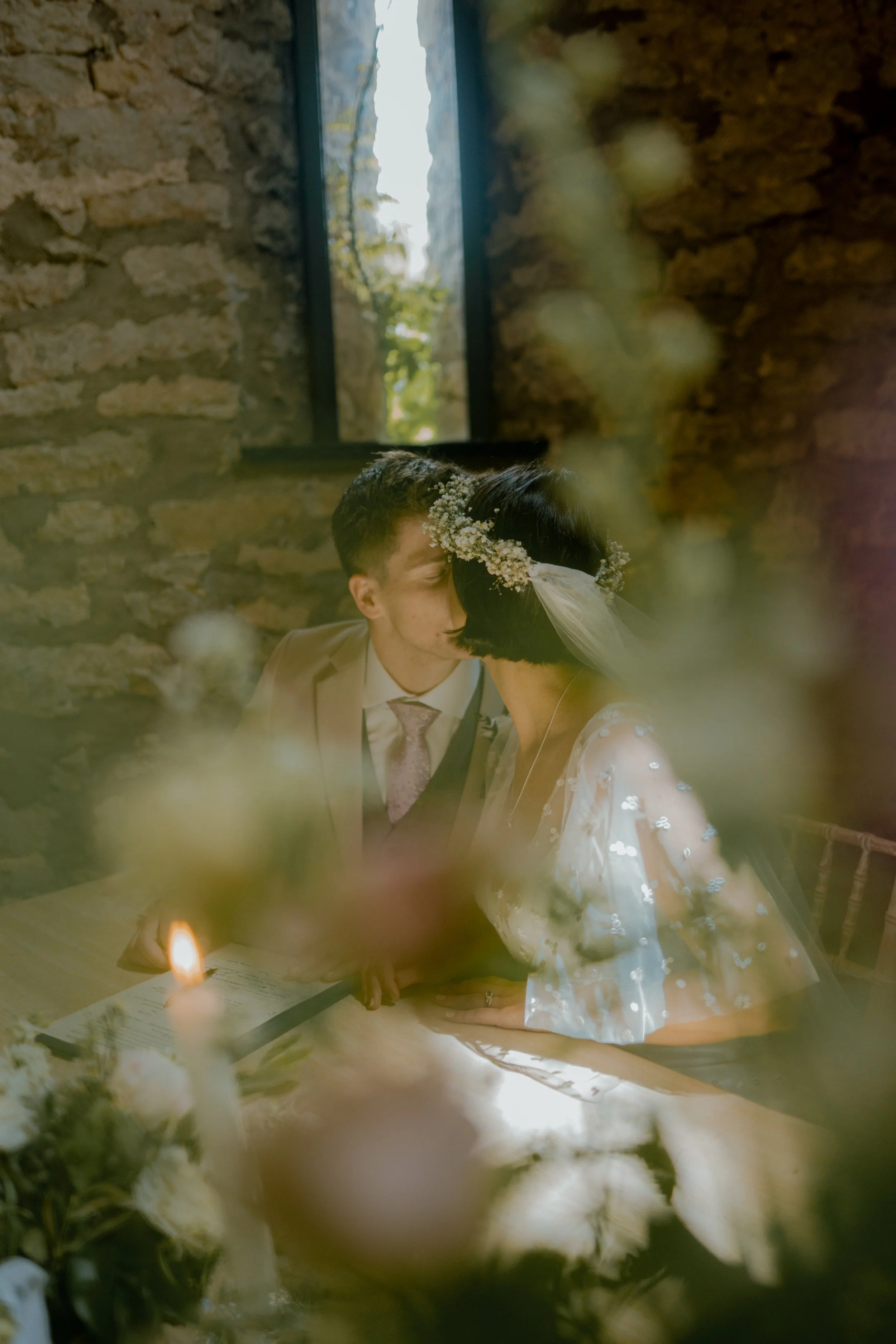 A couple at a wedding ceremony, sitting at a table, sharing a kiss. The woman is wearing a floral headpiece and veil, the man in a beige suit with a pink tie. The scene is softly lit with floral decorations and a candle on the table, with a stone wal