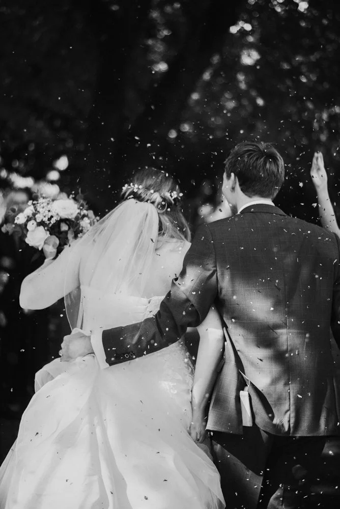 Black and white photo of a bride and groom dancing at their wedding, surrounded by falling confetti and blurred guests, outdoors with trees in the background.