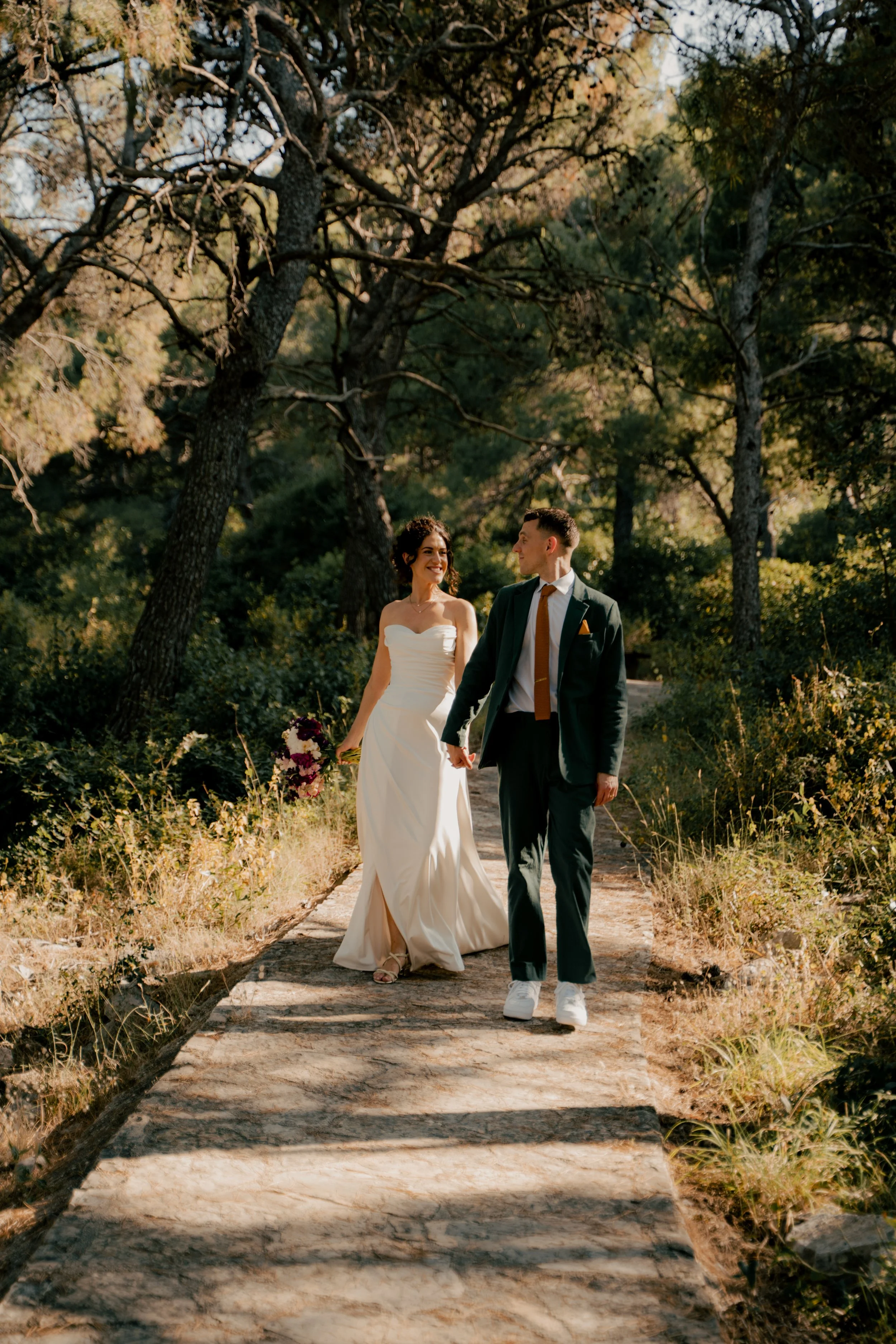 A bride and groom walking hand in hand on a wooded path during sunset, surrounded by trees and greenery.