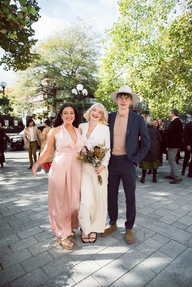 Three young adults dressed in formal attire smiling and posing outdoors, with trees and other people in background.